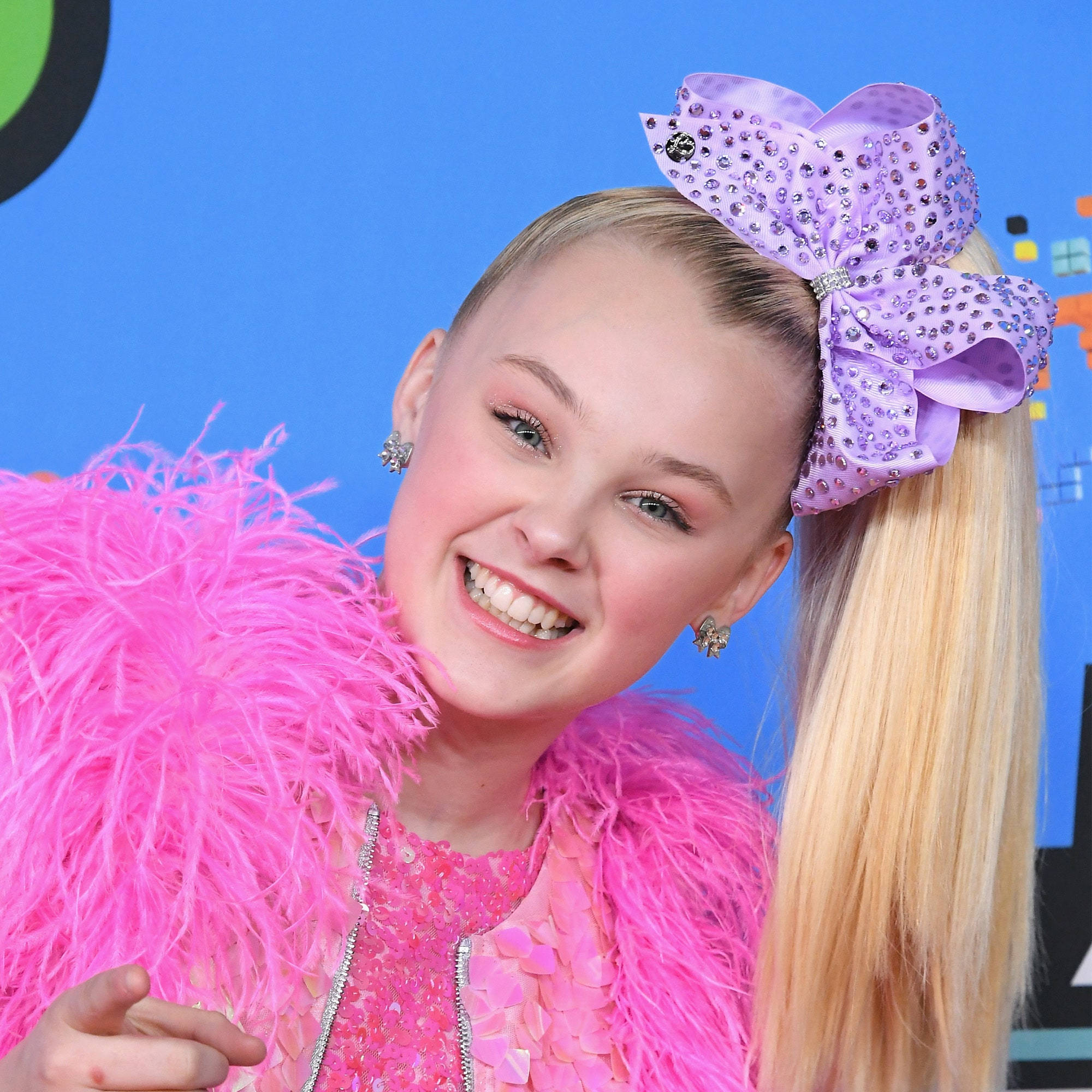 Jojo Siwa 熱情微笑 壁紙 — 一位金髮少女戴著巨大的紫色寶石蝴蝶結，身穿鮮豔的粉紅色羽毛服裝，在藍色背景前燦爛微笑。