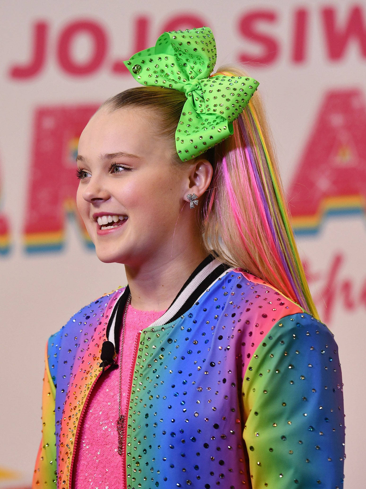 Jojo Siwa 穿彩虹外套 壁紙 — JoJo Siwa 穿著彩虹閃片外套，色彩繽紛的馬尾上繫著一個巨大的綠色蝴蝶結，露出燦爛的笑容。