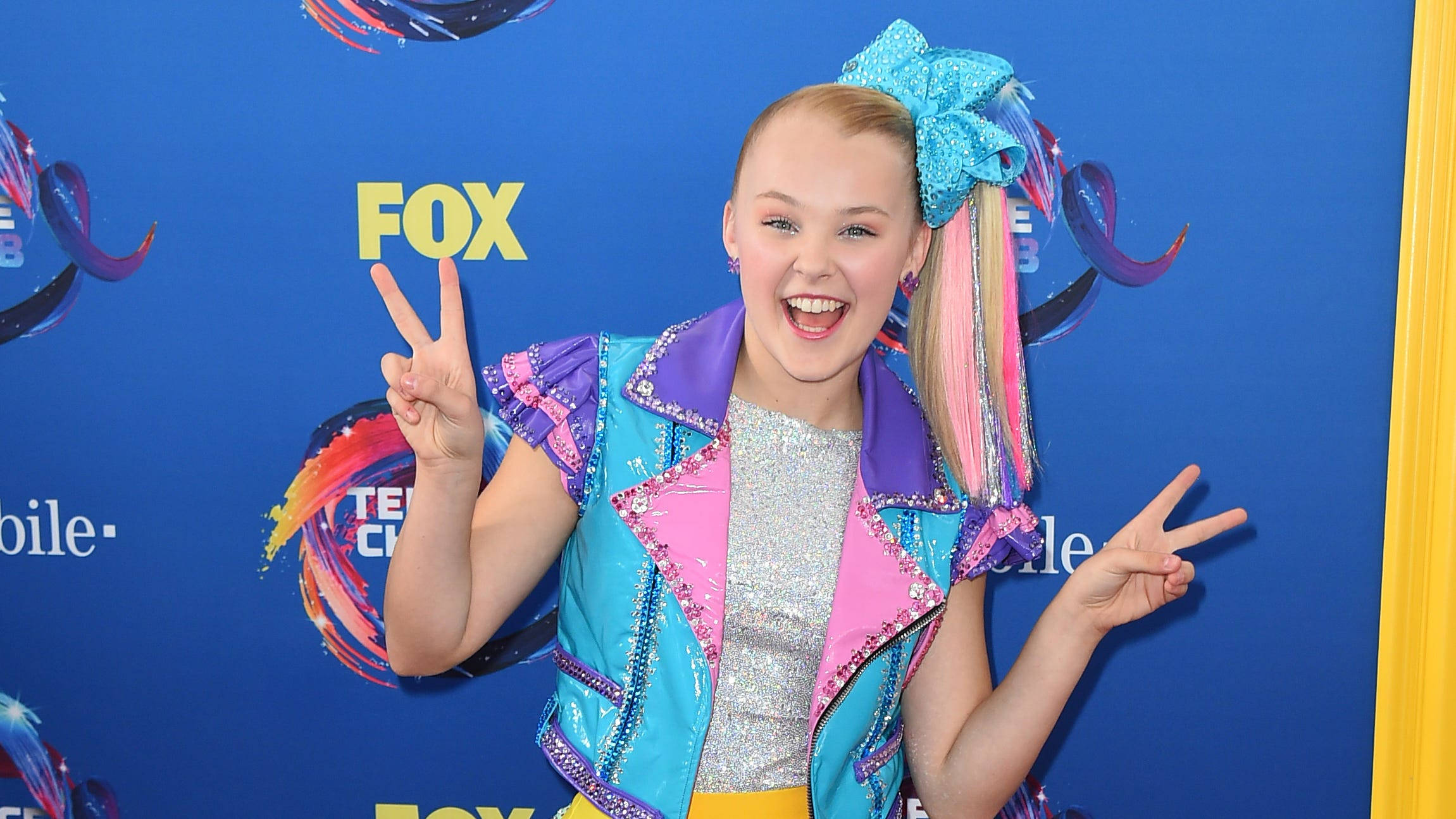 Jojo Siwa 穿著甜美造型 壁紙 — 一個微笑的年輕女孩，戴著一個大藍色蝴蝶結和色彩繽紛的頭髮，在藍色FOX活動背景前做出和平手勢。