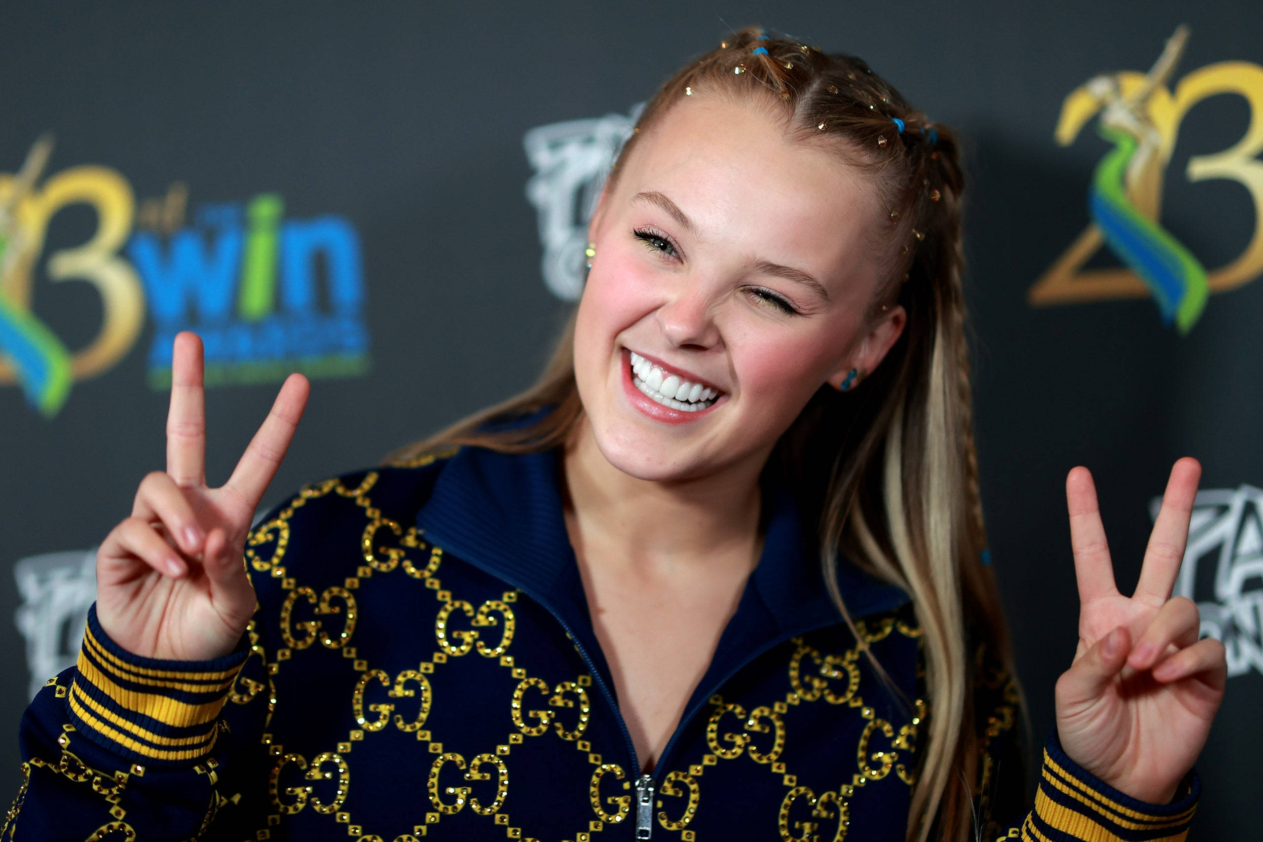 Jojo Siwa 笑容滿面 壁紙 — 一位微笑的年輕女子，編著辮子，在頒獎活動中穿著藍色和金色的Gucci外套，比出和平手勢。