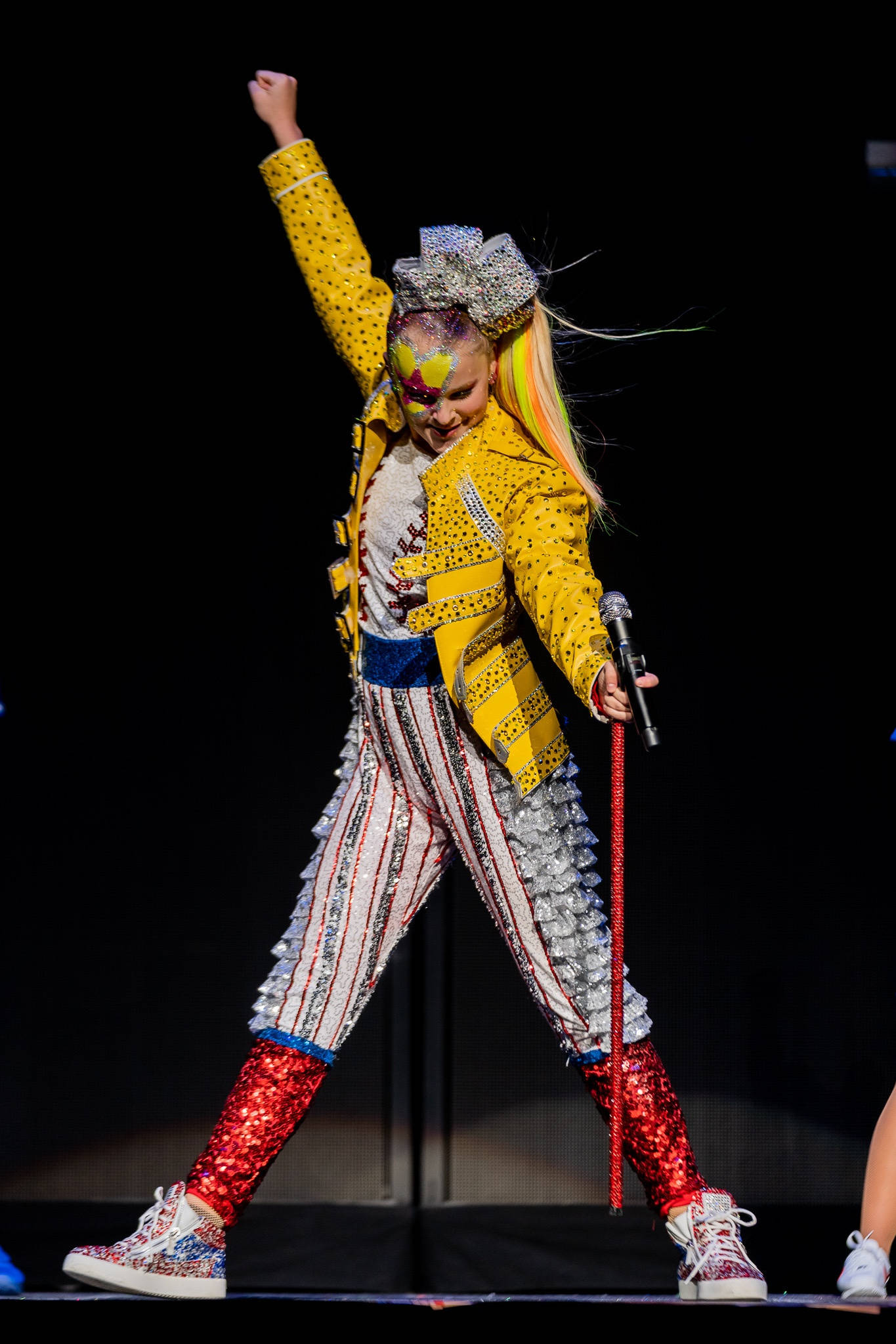Jojo Siwa 舞台演出照 壁紙 — 一位表演者身穿閃亮黃色外套和條紋褲，在舞台上擺出動感姿勢，一隻手臂高舉，手中拿著麥克風。