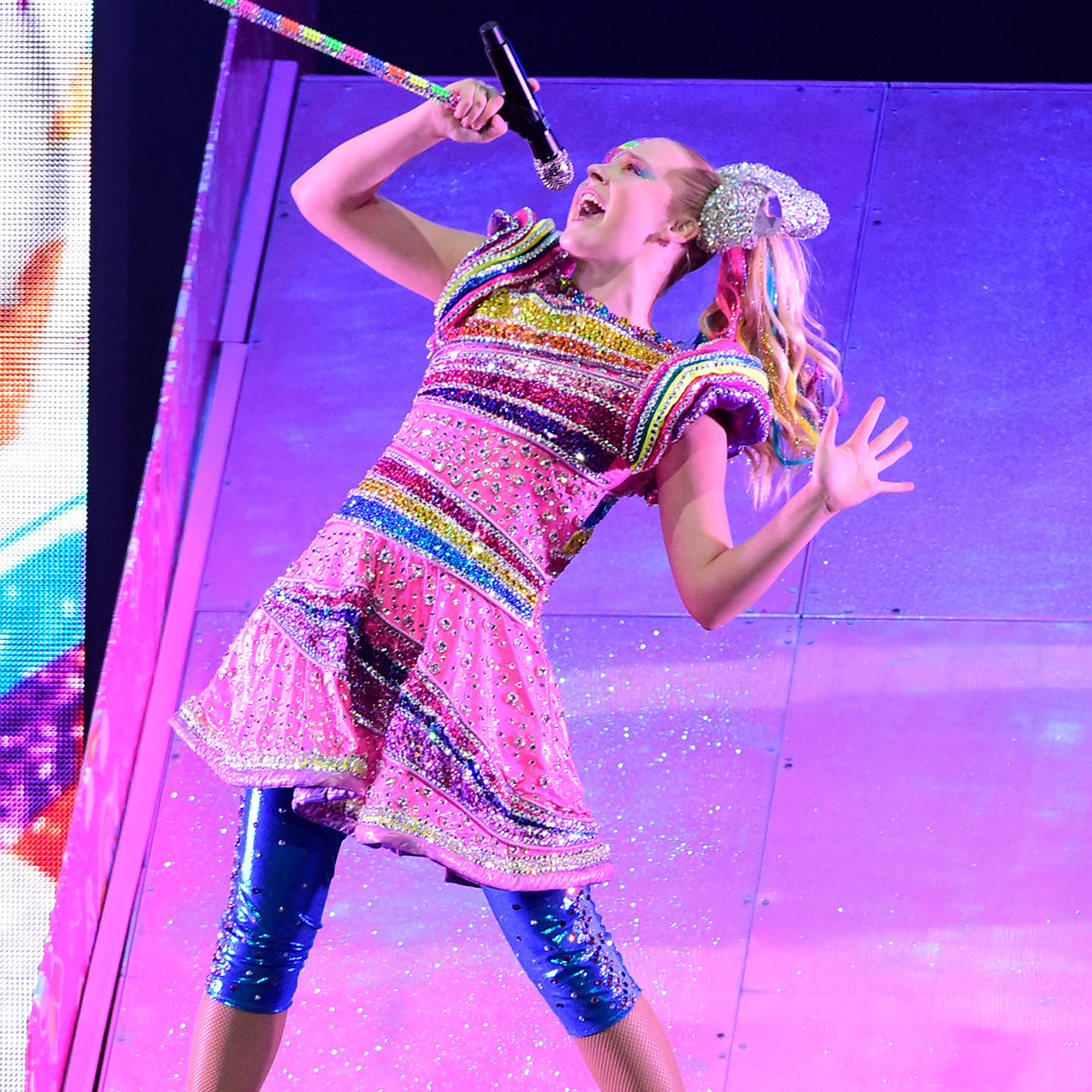 Jojo Siwa 舞台演唱 壁紙 — 一位充滿活力的表演者身穿閃閃發光的彩虹服裝，在紫色燈光的舞台上激情演唱，火花四濺。