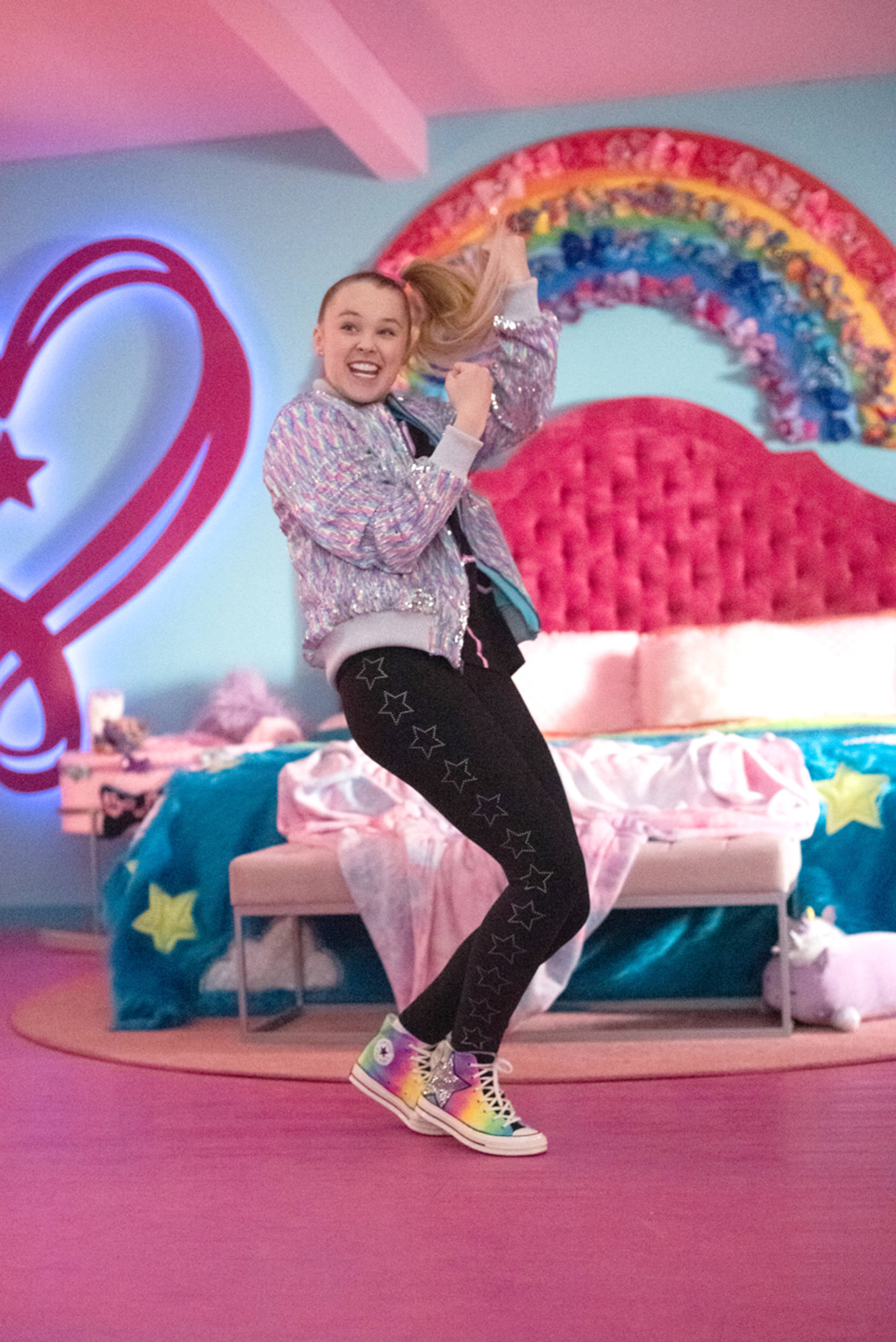 Jojo Siwa 舞蹈隨拍 壁紙 — 一個快樂的女孩穿著閃亮外套和星星緊身褲，在充滿活力的粉紅色房間裡，配著彩虹床頭板，充滿活力地跳舞。