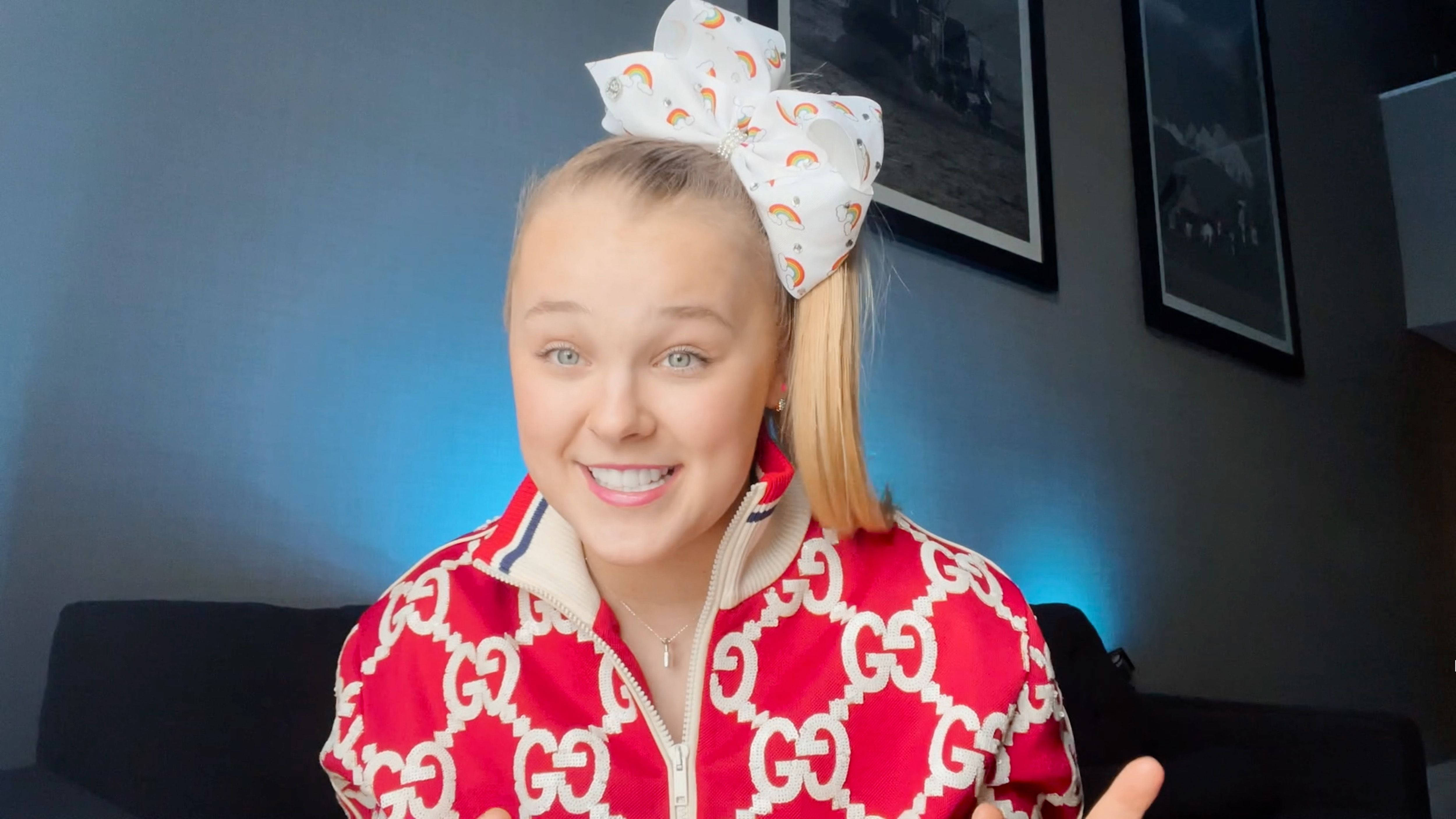 Jojo Siwa 身穿 Gucci 套裝 壁紙 — 一位開朗的年輕女性，金髮紮成馬尾，戴著一個大彩虹蝴蝶結，身穿紅色GG圖案外套，在室內對著鏡頭微笑。