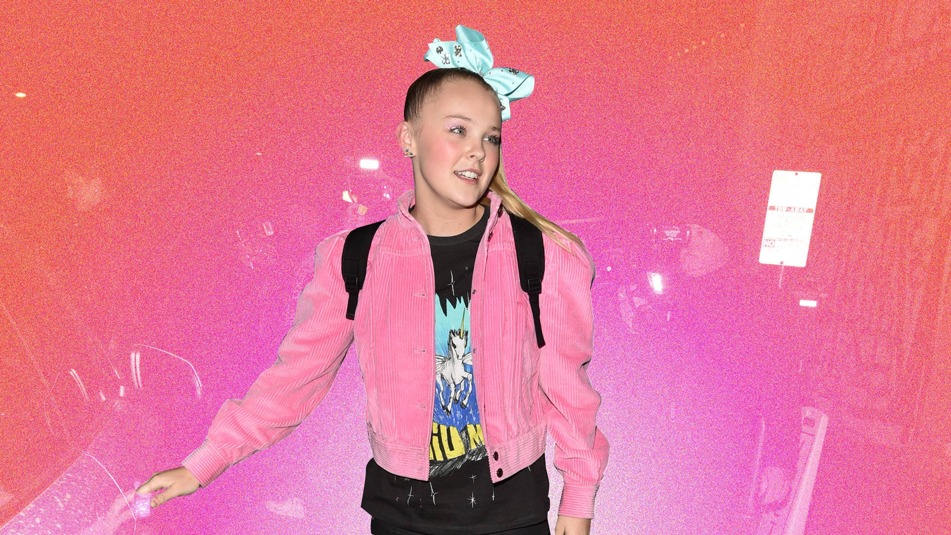 Jojo Siwa 身穿粉紅針織開衫 壁紙 — 一個年輕女孩戴著一個大藍色蝴蝶結，穿著粉紅色外套，在充滿活力的粉紅色和橙色漸變牆前微笑。