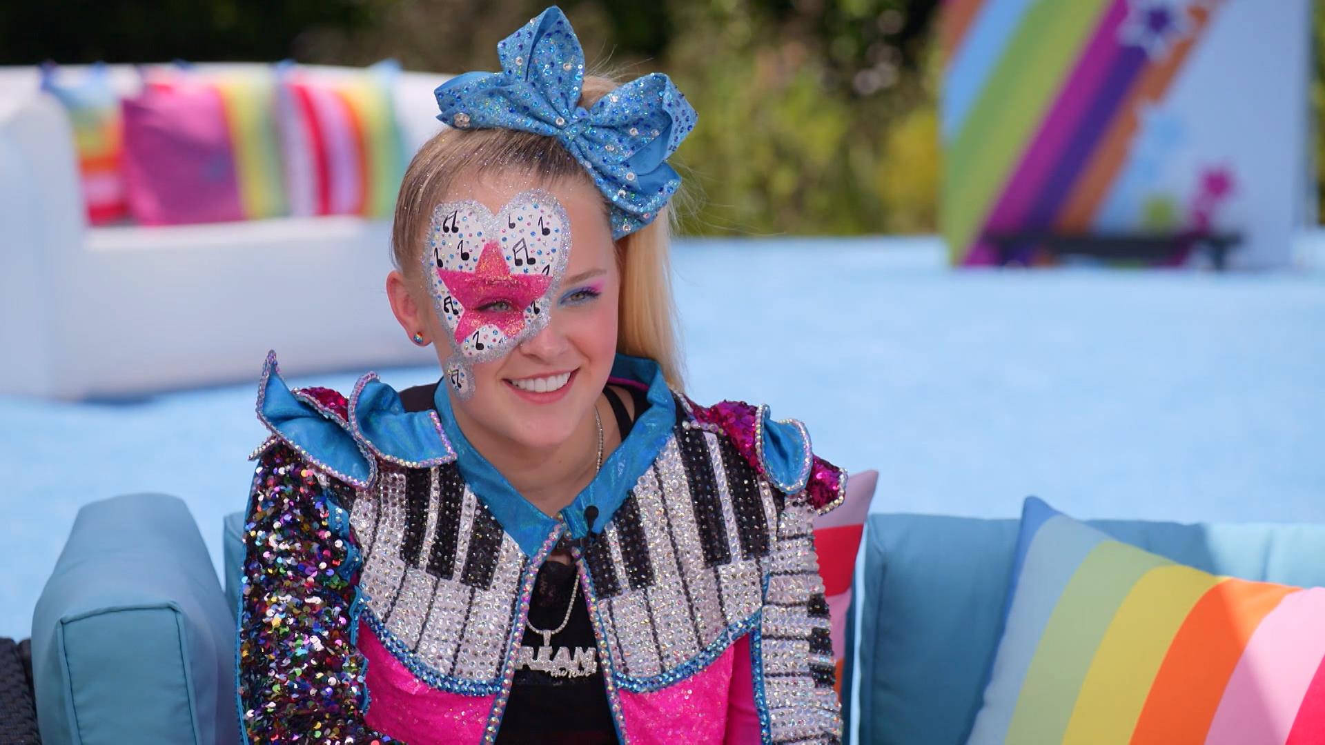 Jojo Siwa 面部彩繪 壁紙 — 一個微笑的女孩穿著閃閃發光的鋼琴鍵外套和藍色蝴蝶結，坐在帶有彩虹枕頭的彩色沙發上。