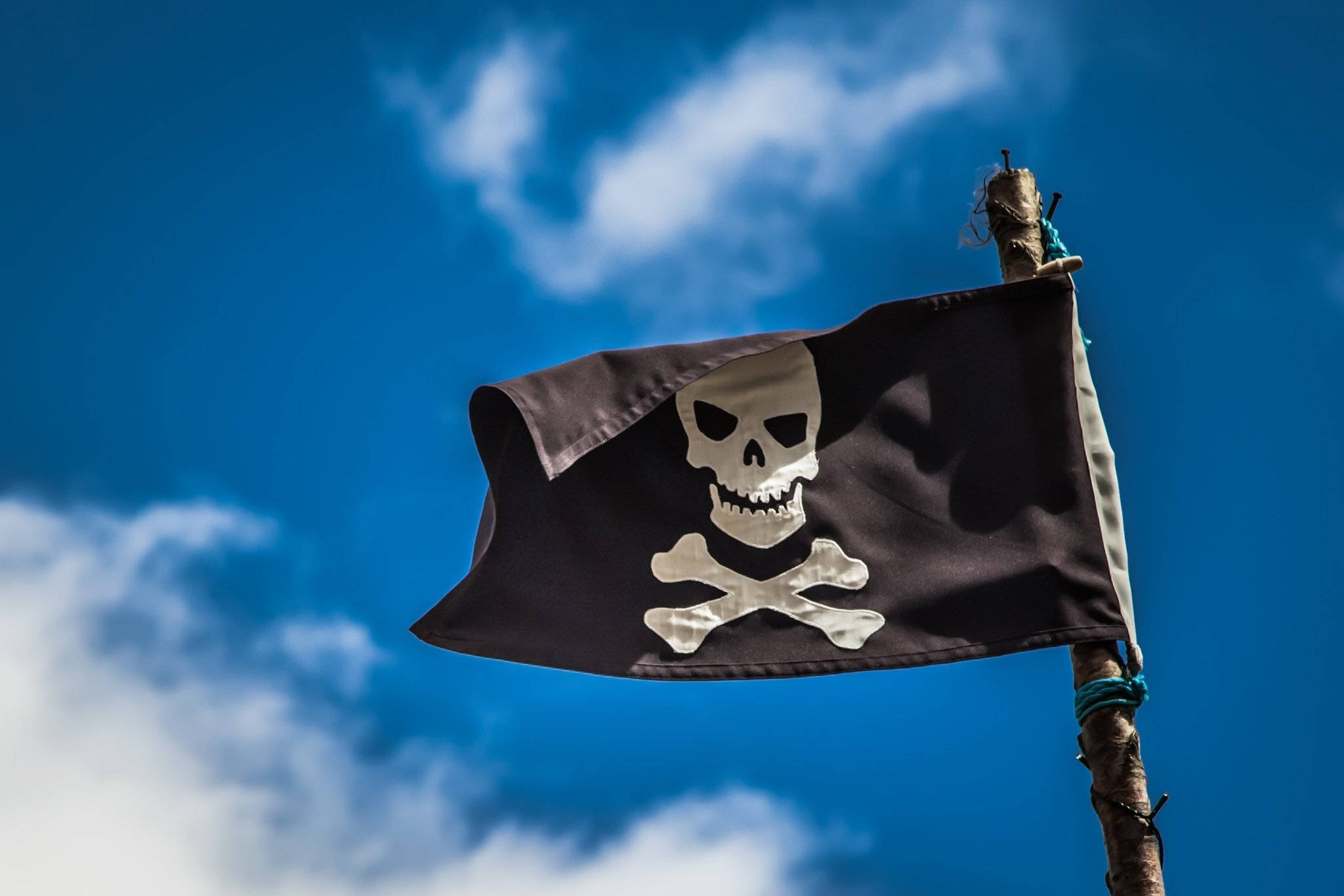賊船旗（Jolly Roger） 壁紙 — 一面破爛的黑色海盜旗，上面有白色骷髏頭和交叉骨頭，在藍天白雲中飄揚。