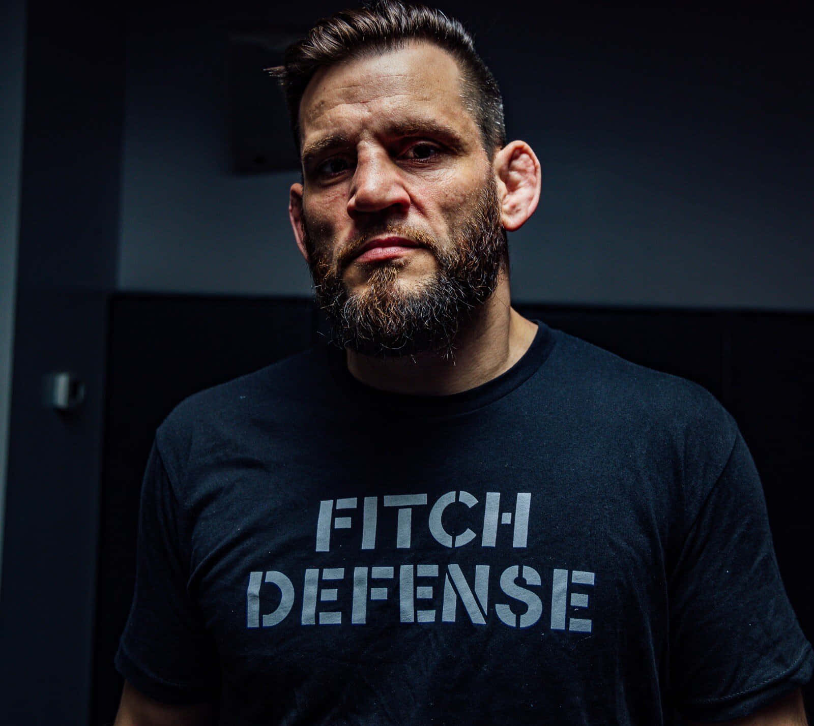 Jon Fitch 自衛教練形象 壁紙 — 一個留鬍鬚的男人，身穿黑色 'FITCH DEFENSE' T恤，在昏暗的房間裏凝視著鏡頭。