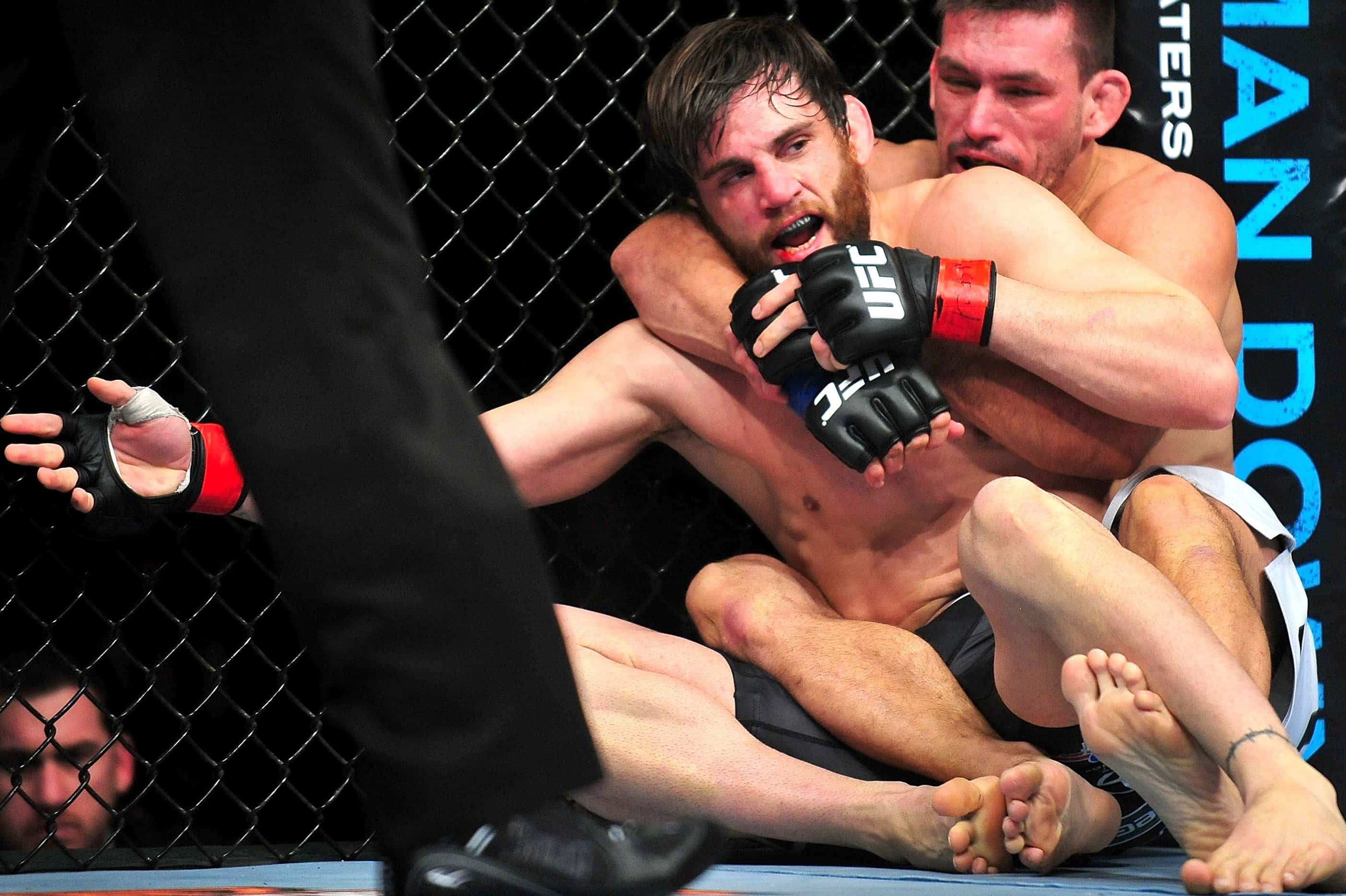 Jon Fitch賽中被壓角 壁紙 — UFC比賽中，兩名拳手在黑色鐵絲網籠內的藍色墊子上激烈纏鬥。