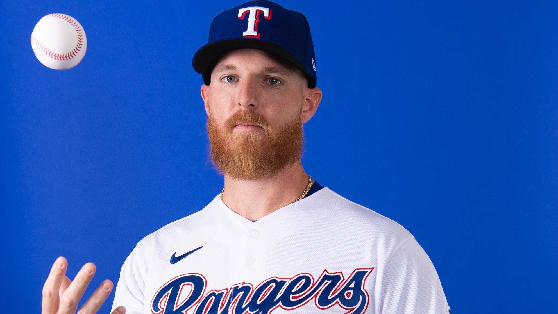 Jon Gray 玩球 壁紙 — 一名留鬍鬚的棒球運動員身穿 Rangers 球衣，在純藍色攝影棚背景下投擲棒球。