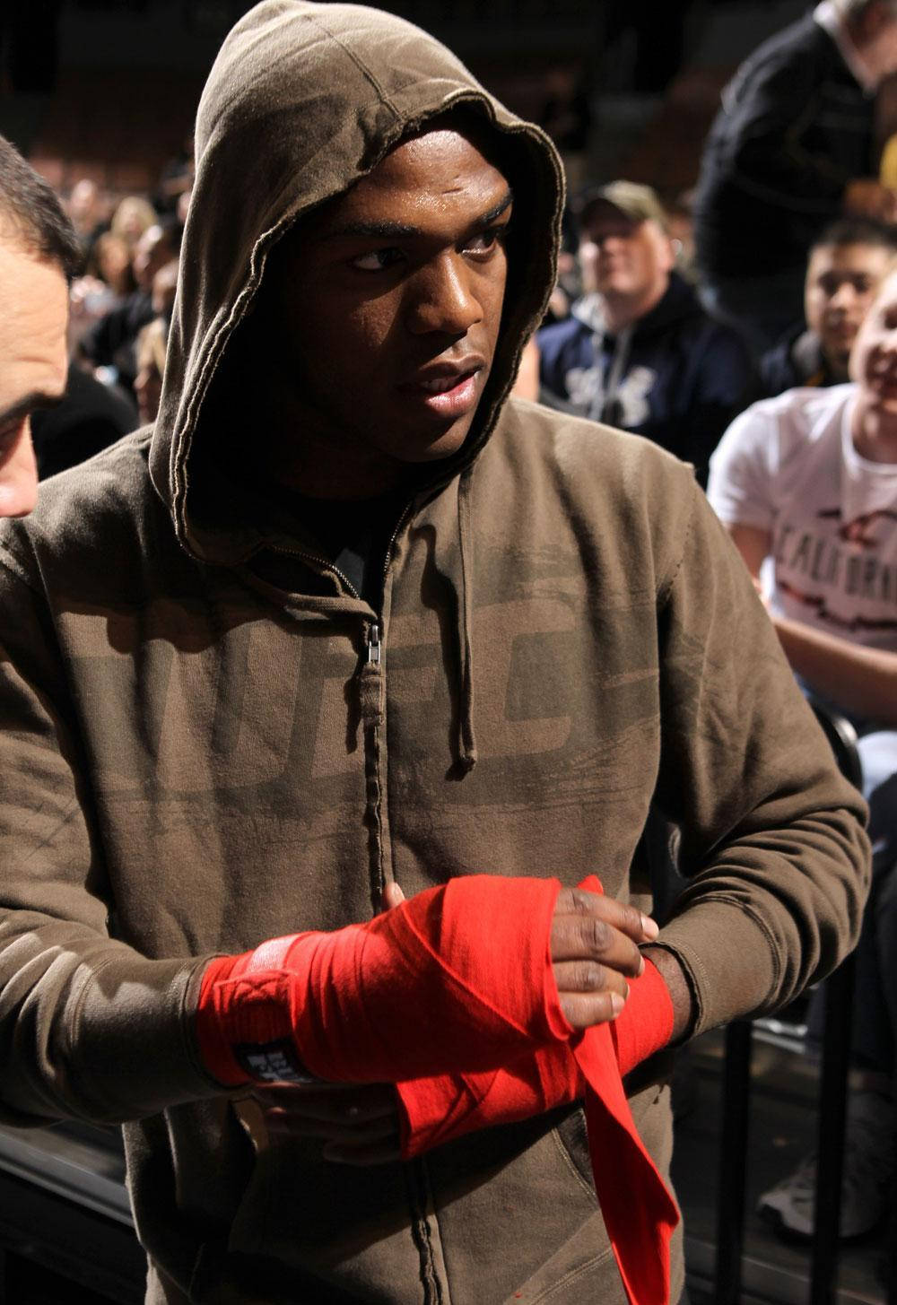 Jon Jones 包手前的準備 壁紙 — 一位專注的戰士穿著棕色連帽衫，在競技場燈光下比賽前用亮紅色膠帶纏繞他的手。