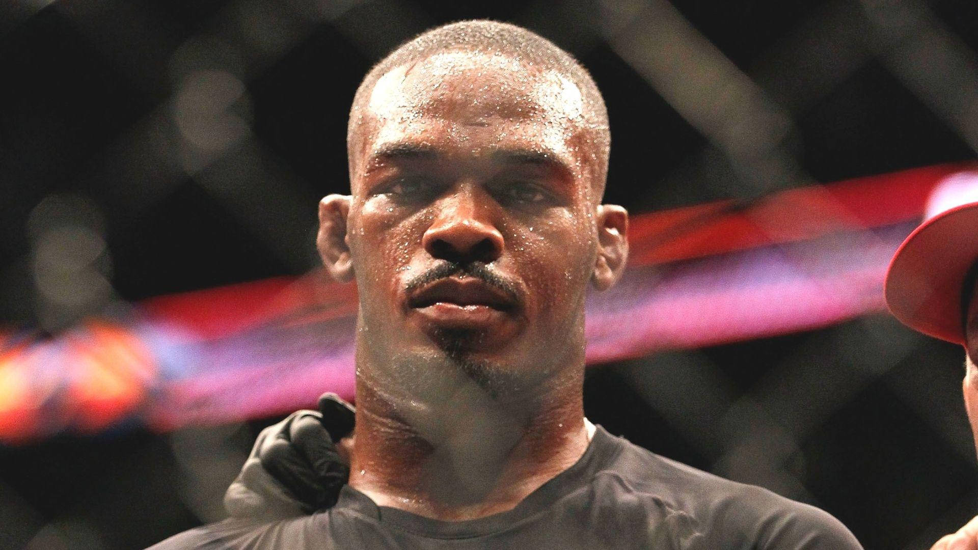 汗水淋漓的 Jon Jones 壁紙 — 一個滿身汗水、意志堅定的拳手在昏暗的MMA籠中，背後有模糊的紅光，眼神銳利地凝視著。
