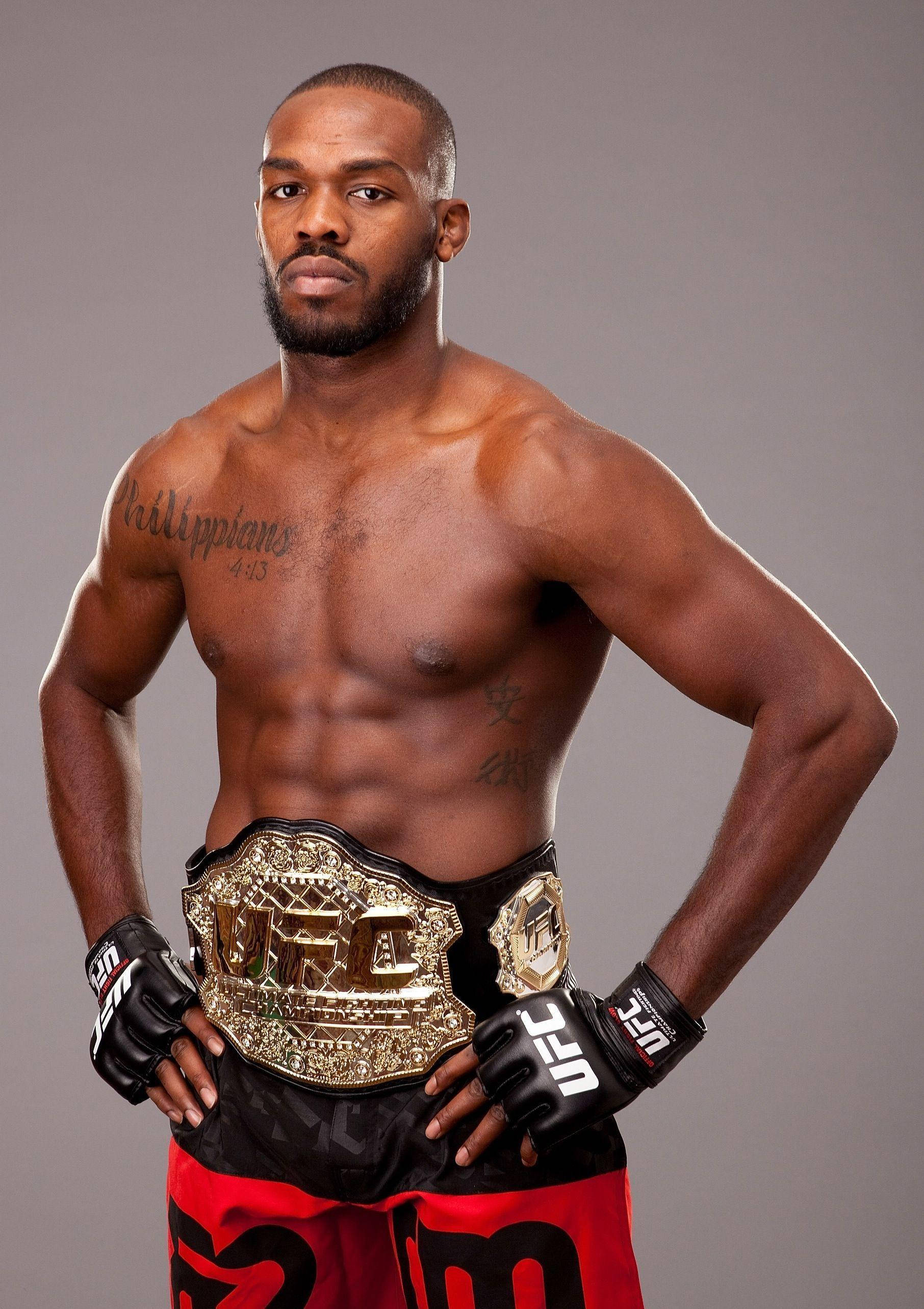 Jon Jones 正式肖像照 壁紙 — 一位肌肉發達、有紋身的UFC拳手自信地站立，手持冠軍腰帶，背景是純灰色攝影棚。
