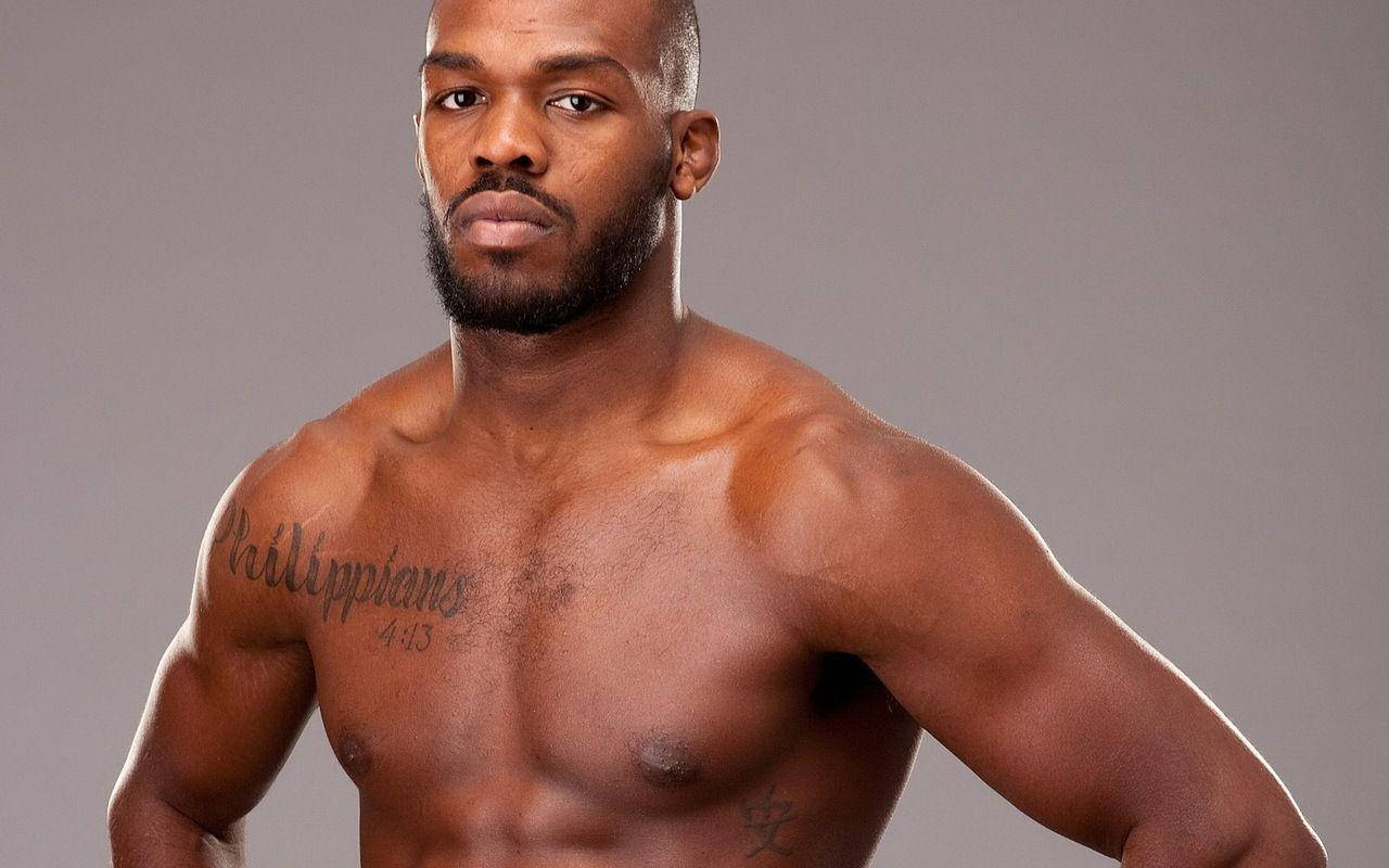 Jon Jones 細節側影照 壁紙 — 一個肌肉發達、留鬍鬚、有紋身的黑人男子，在純灰色工作室背景下，強烈地凝視著鏡頭。