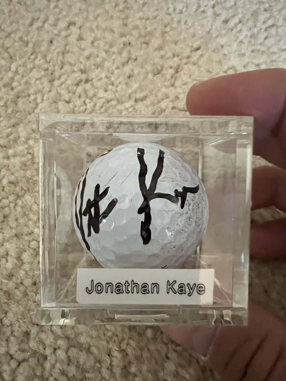 Jonathan Kaye親筆簽名高爾夫球 壁紙 — 一隻手拿著一個透明亞加力展示盒，內有簽名高爾夫球，背景是米色地毯。