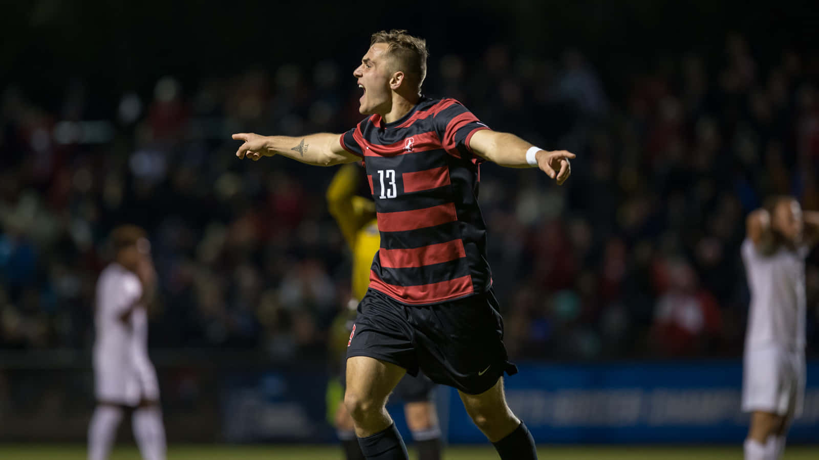 Jordan Morris 2015 男子足球隊賽事影像 壁紙 — 一名身穿紅黑間條球衣的足球員在球場燈光下張開雙臂慶祝入球。