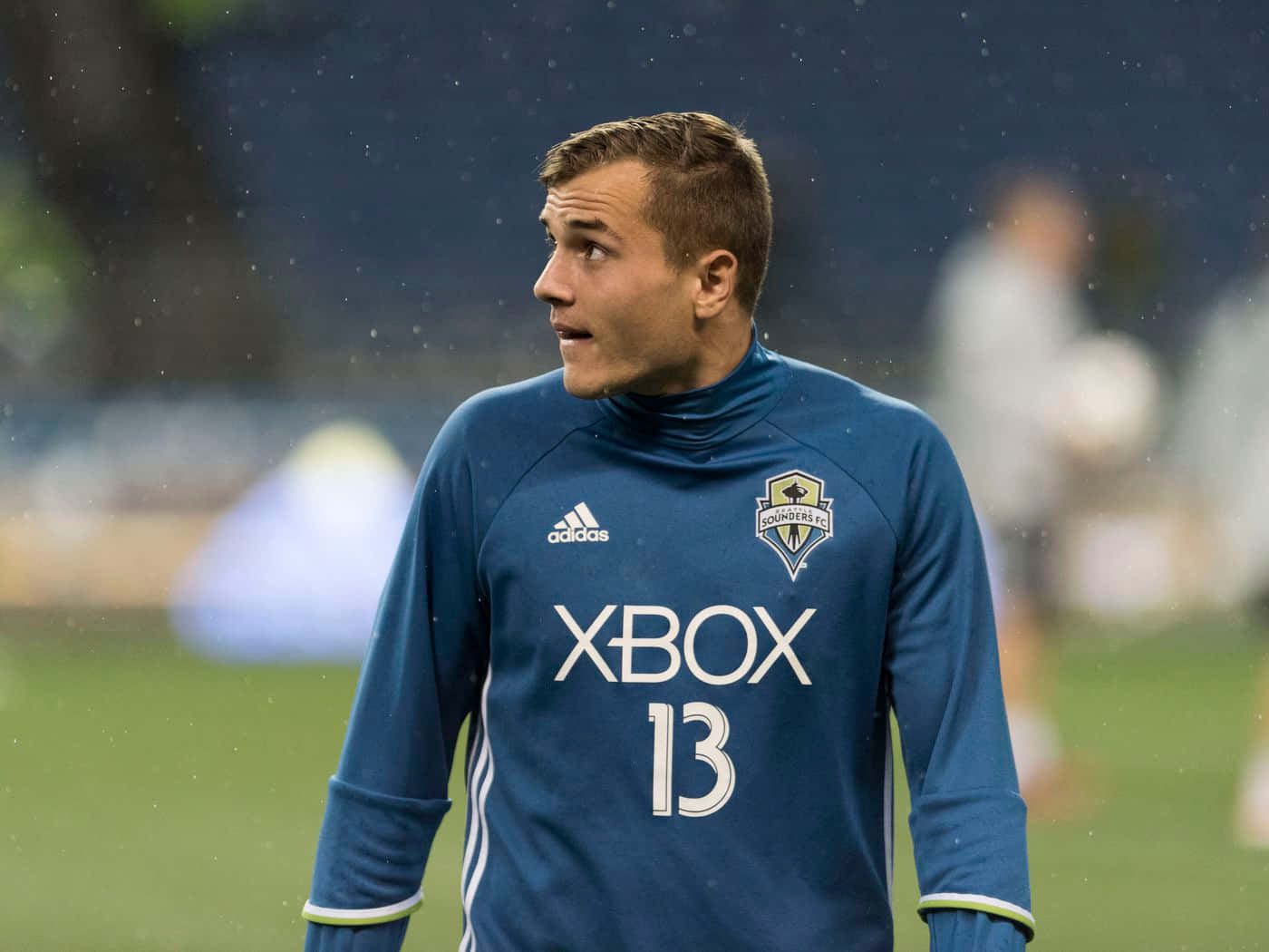 Jordan Morris 側面角度美照 壁紙 — 一名身穿帶有Xbox標誌的藍色訓練球衣的足球運動員站在被雨淋濕的場地上，視線離開鏡頭。