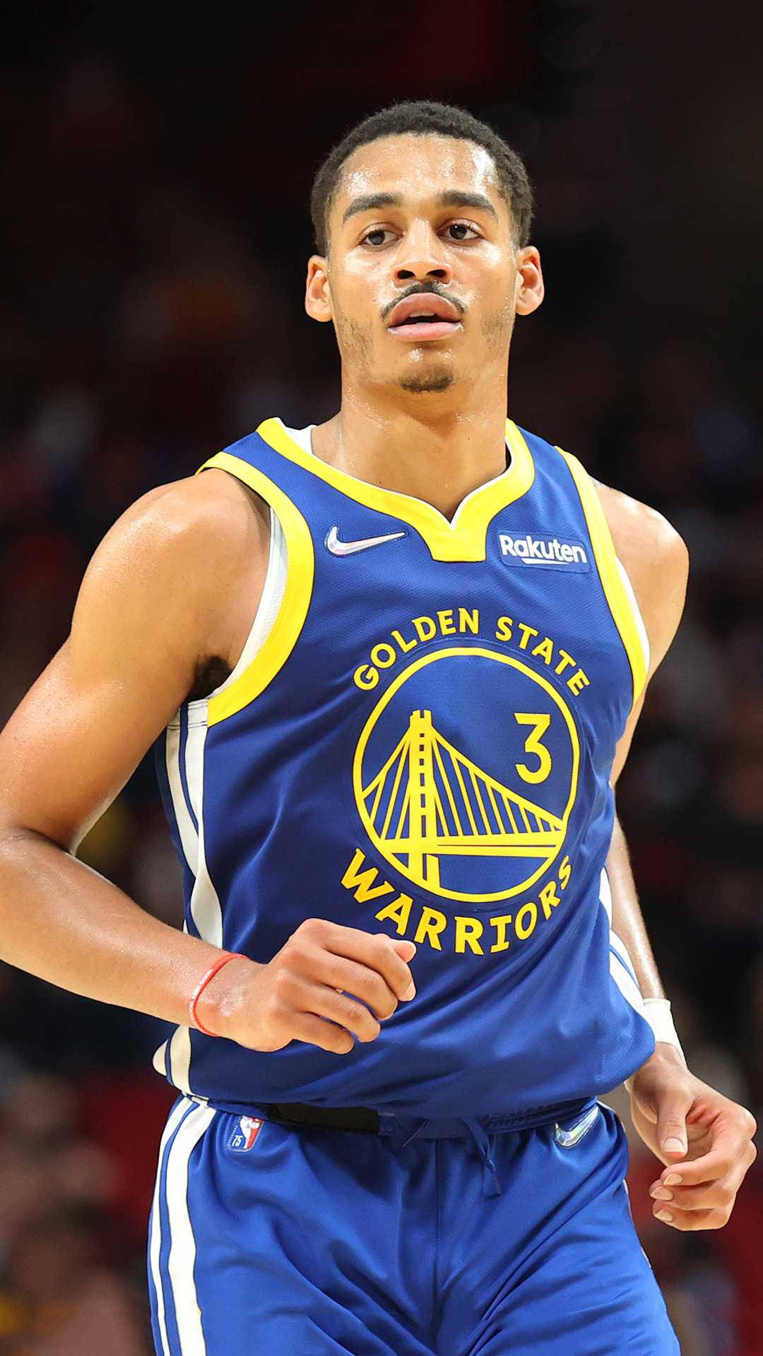 Jordan Poole賽中奔跑 壁紙 — 一位專注的金州勇士籃球運動員穿著藍黃球衣在場館燈光下比賽中奔跑。