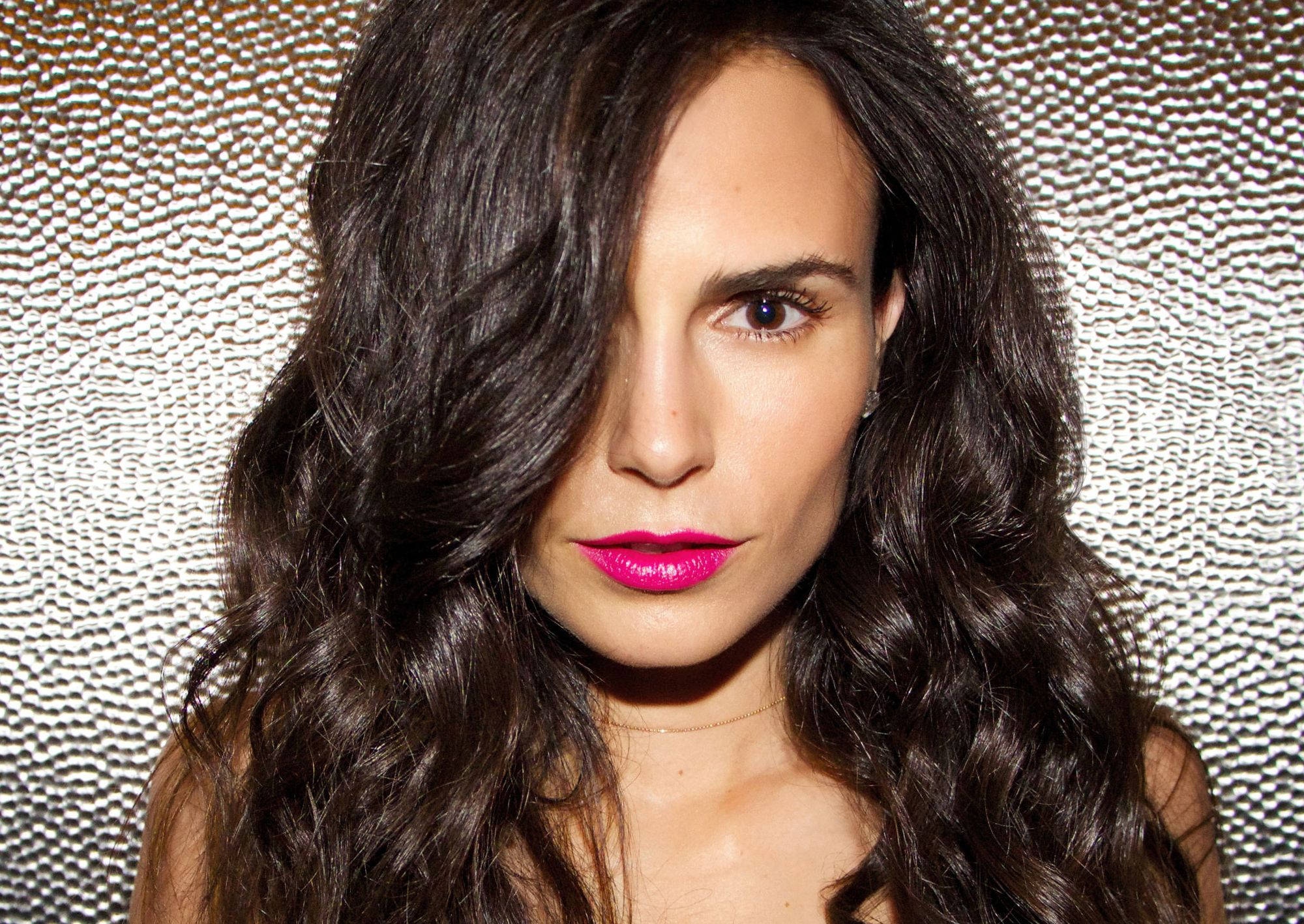 Jordana Brewster 粉紅唇妝 壁紙 — 一位擁有深色波浪頭髮和大膽粉紅嘴唇的女性，在帶有紋理的銀色背景下強烈凝視。
