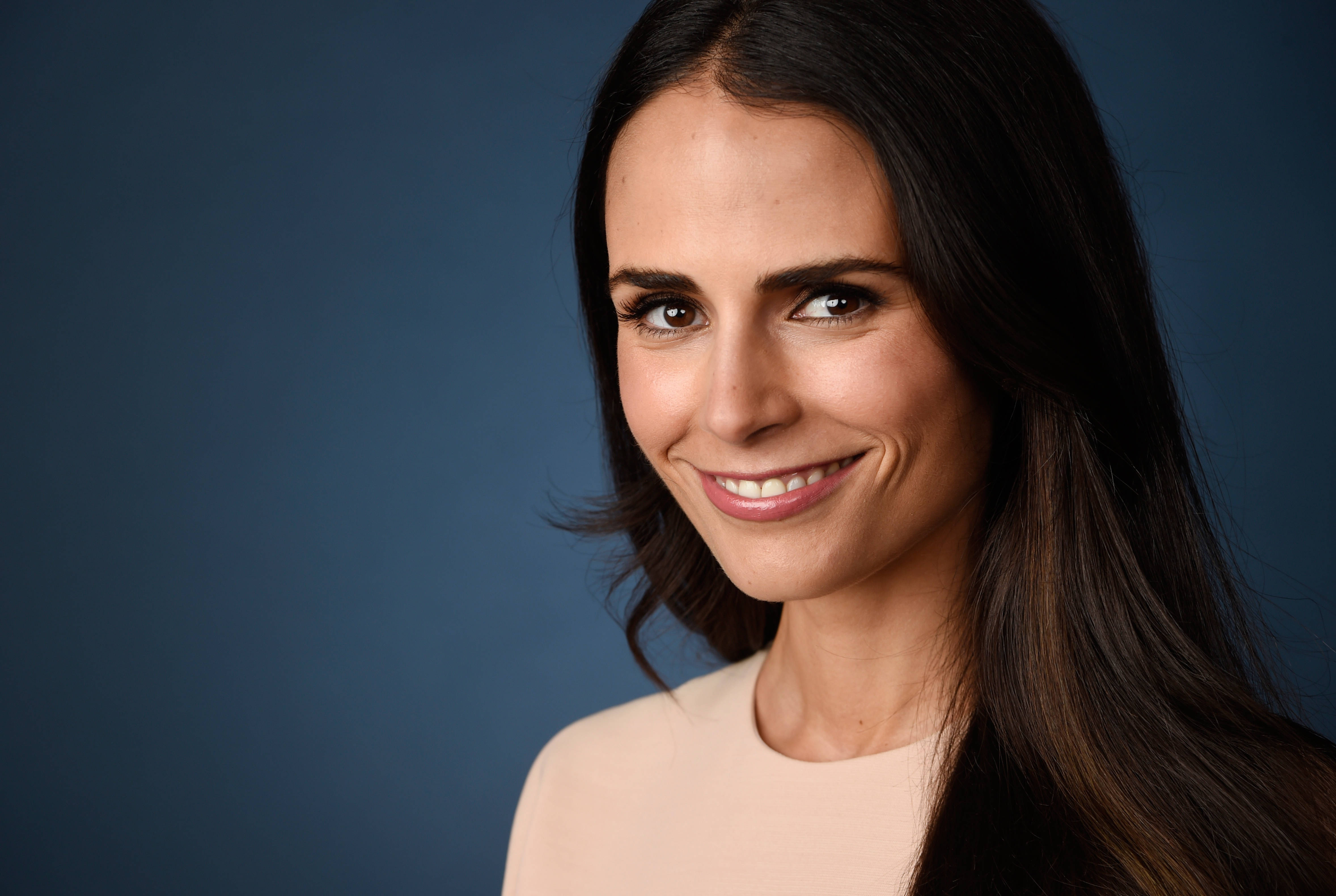 Jordana Brewster 深藍背景微笑特寫 壁紙 — 一位擁有長黑髮的女性在純藍色攝影棚背景前溫暖地微笑。