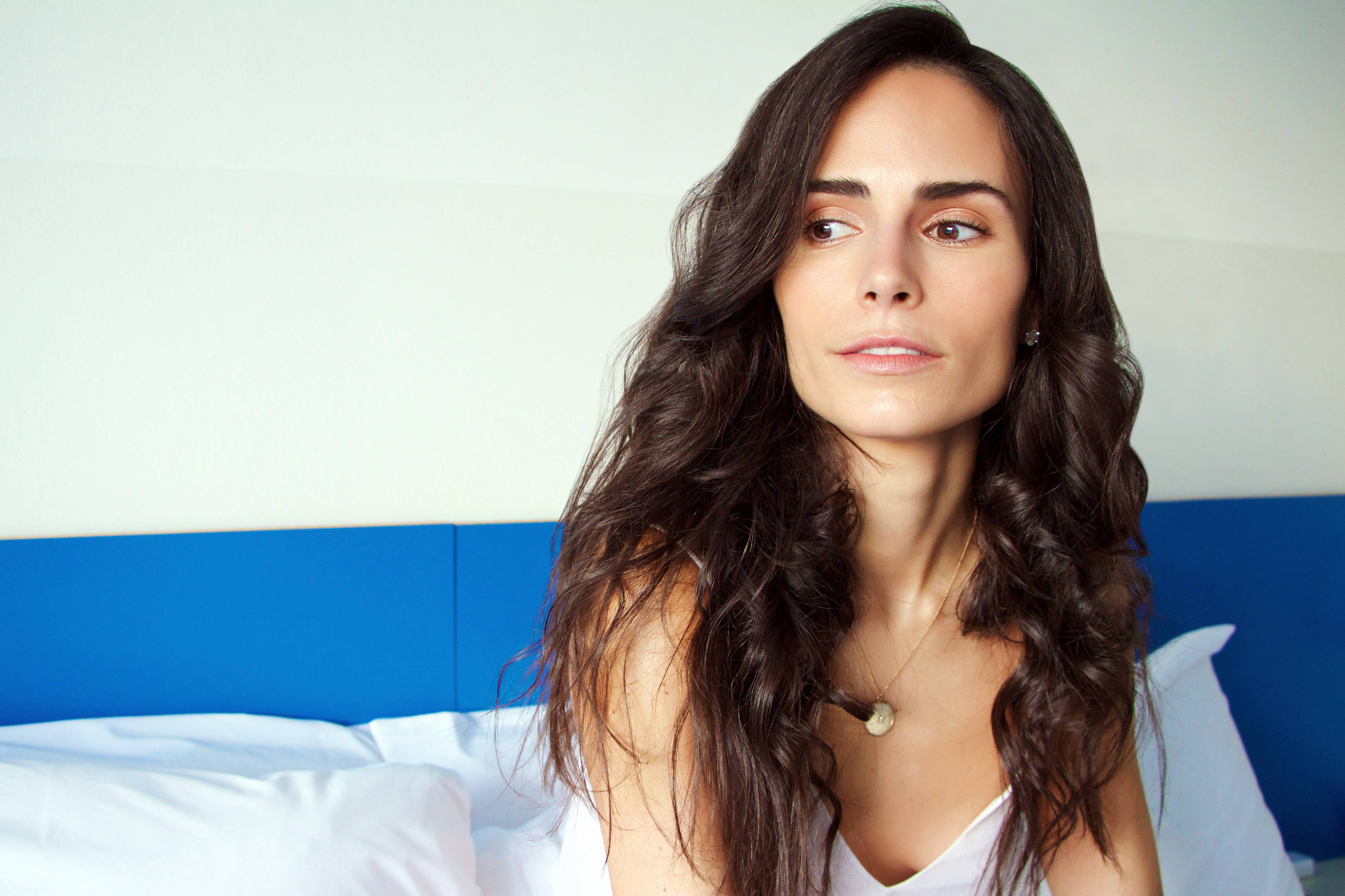 Jordana Brewster 臥室拍攝 壁紙 — 一位長著深色波浪長髮的女性坐在一張有白色床單和藍色床頭板的床上，若有所思地望向遠方。