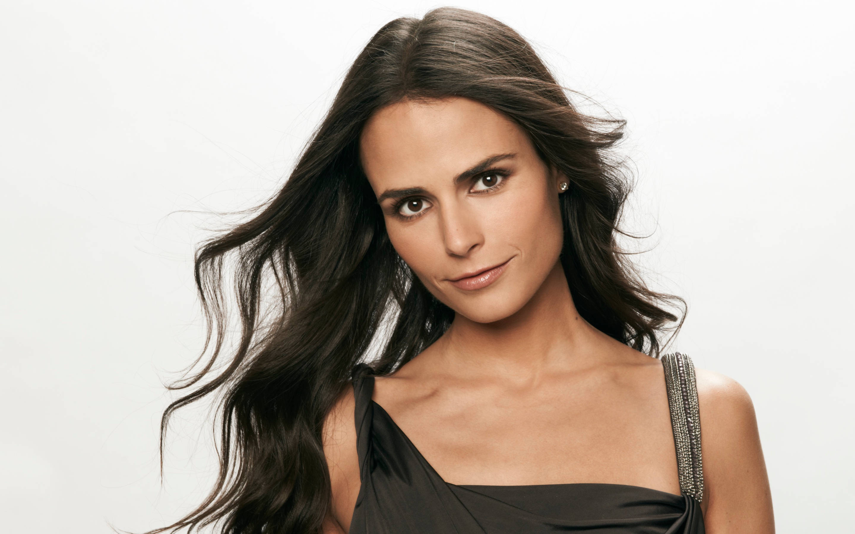 Jordana Brewster 浪漫捲髮造型 壁紙 — 一位長黑髮、帶著微妙微笑的女性，在純白背景前穿著黑色連衣裙。