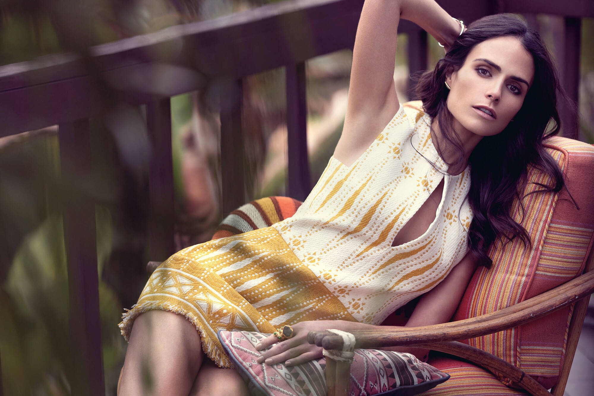 Jordana Brewster 黃色迷你禮服 壁紙 — 一位身穿黃色圖案連衣裙的女性優雅地躺在被綠植環繞的門廊木椅上。