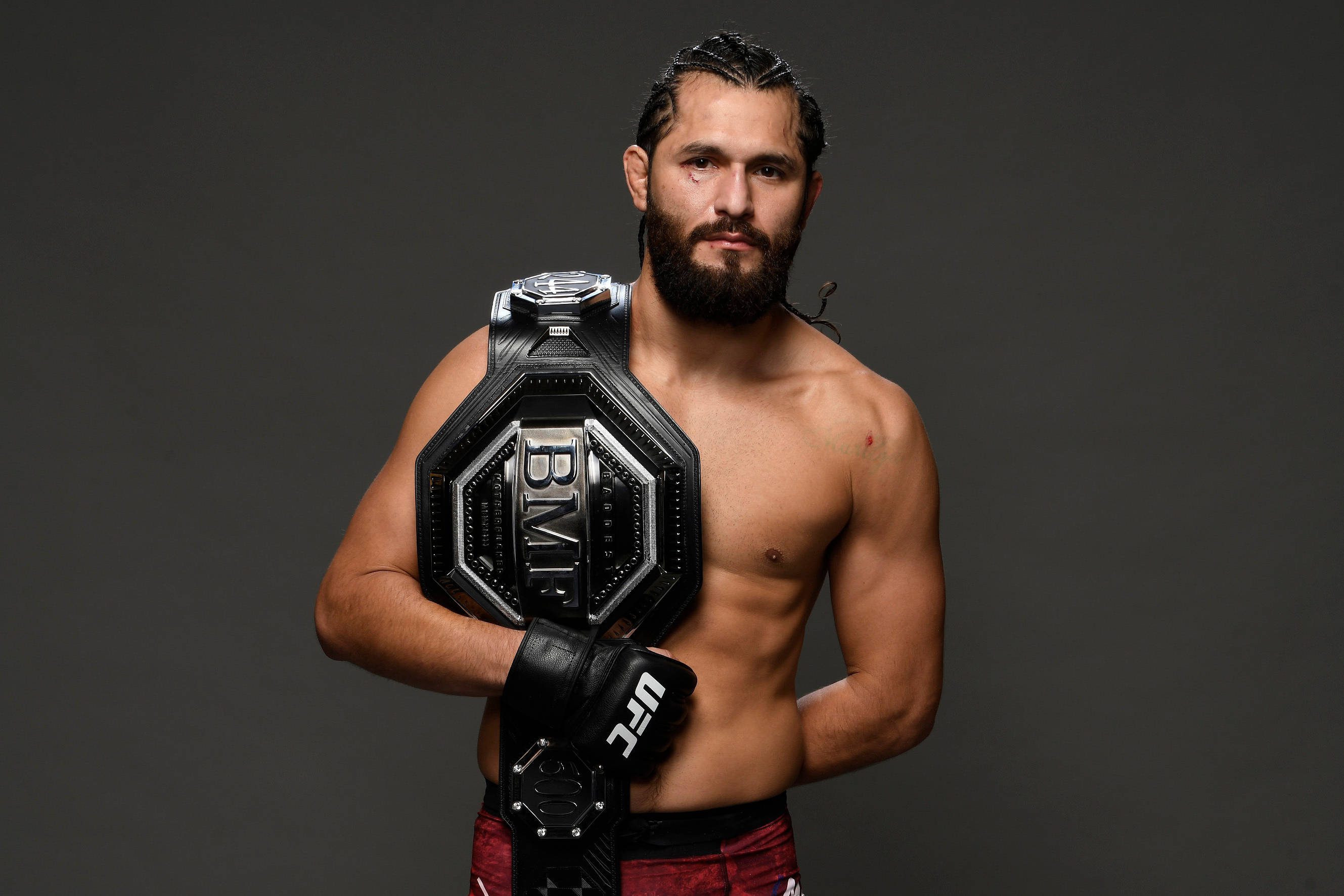 Jorge Masvidal 肩扛腰帶 壁紙 — 一位留鬍子的肌肉發達拳手，在深灰色工作室背景前手持UFC冠軍腰帶。