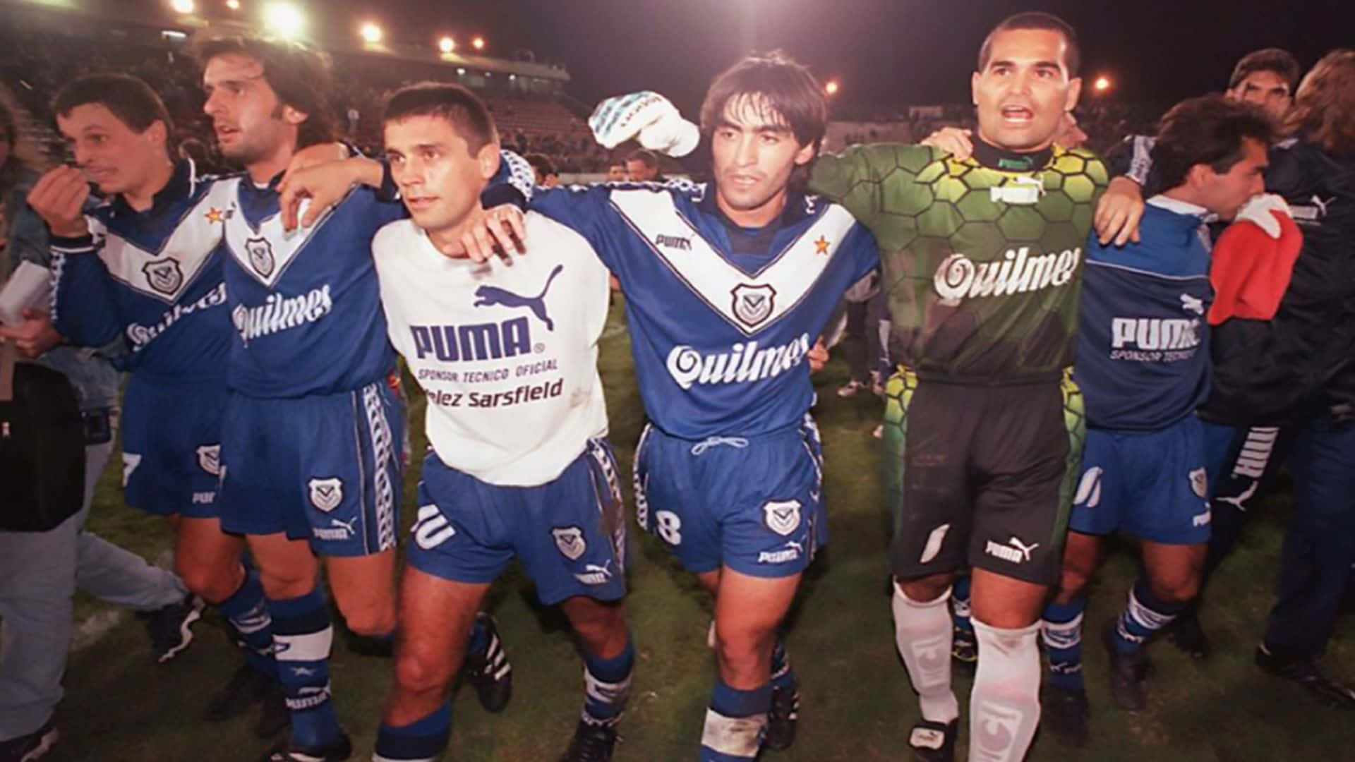 José Luis Chilavert 為Club Atlético Vélez Sarsfield效力時的經典姿勢 壁紙 — 身穿藍白球衣的足球員在夜間球場燈光下的草地上一起慶祝。