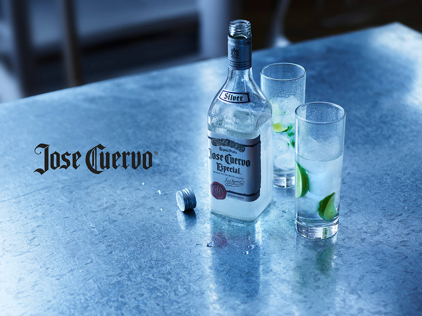 Jose Cuervo 冰鎮雞尾酒 壁紙 — 一瓶冰凍的Jose Cuervo Silver龍舌蘭酒和兩個帶有青檸角的玻璃杯放在涼爽的金屬吧檯面上。
