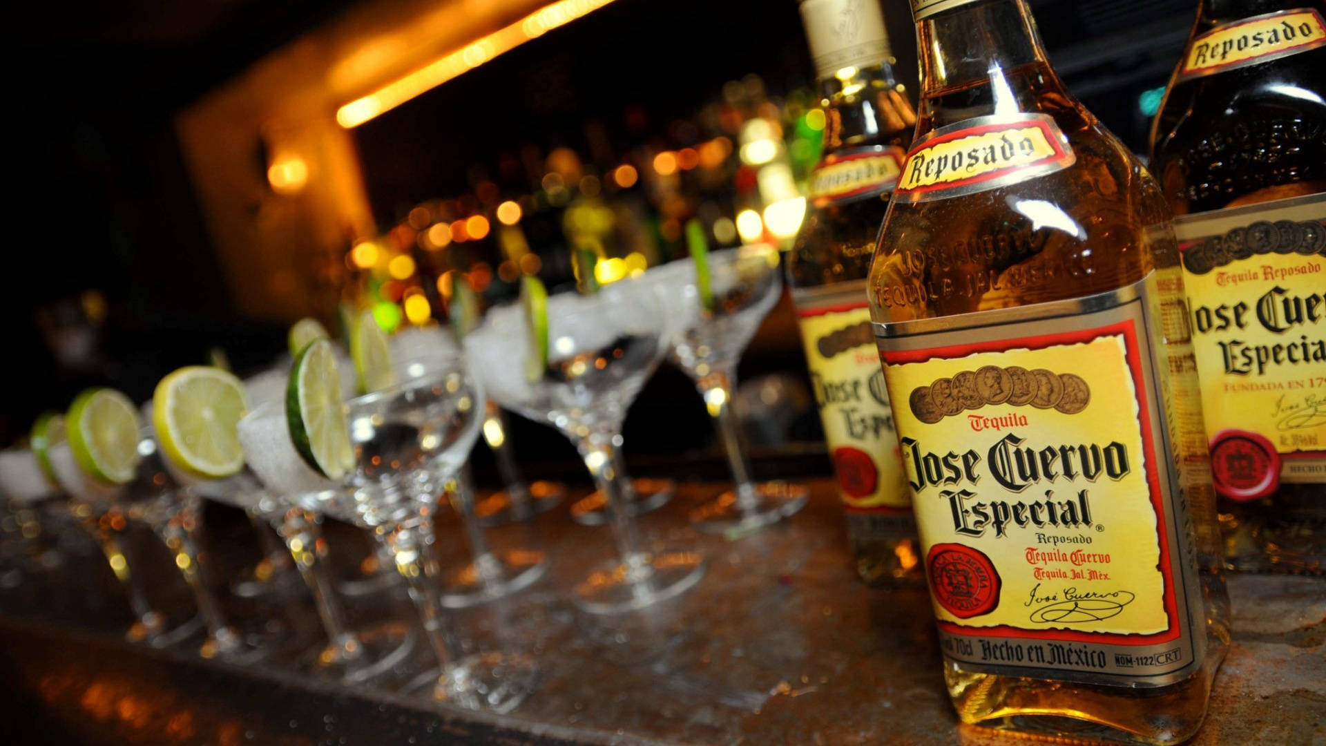 Jose Cuervo Especial 蒸餾酒與檸檬杯 壁紙 — 一排排龍舌蘭酒瓶和以青檸裝飾的瑪格麗特酒杯在昏暗的休息室中，在溫暖的酒吧燈光下閃閃發光。