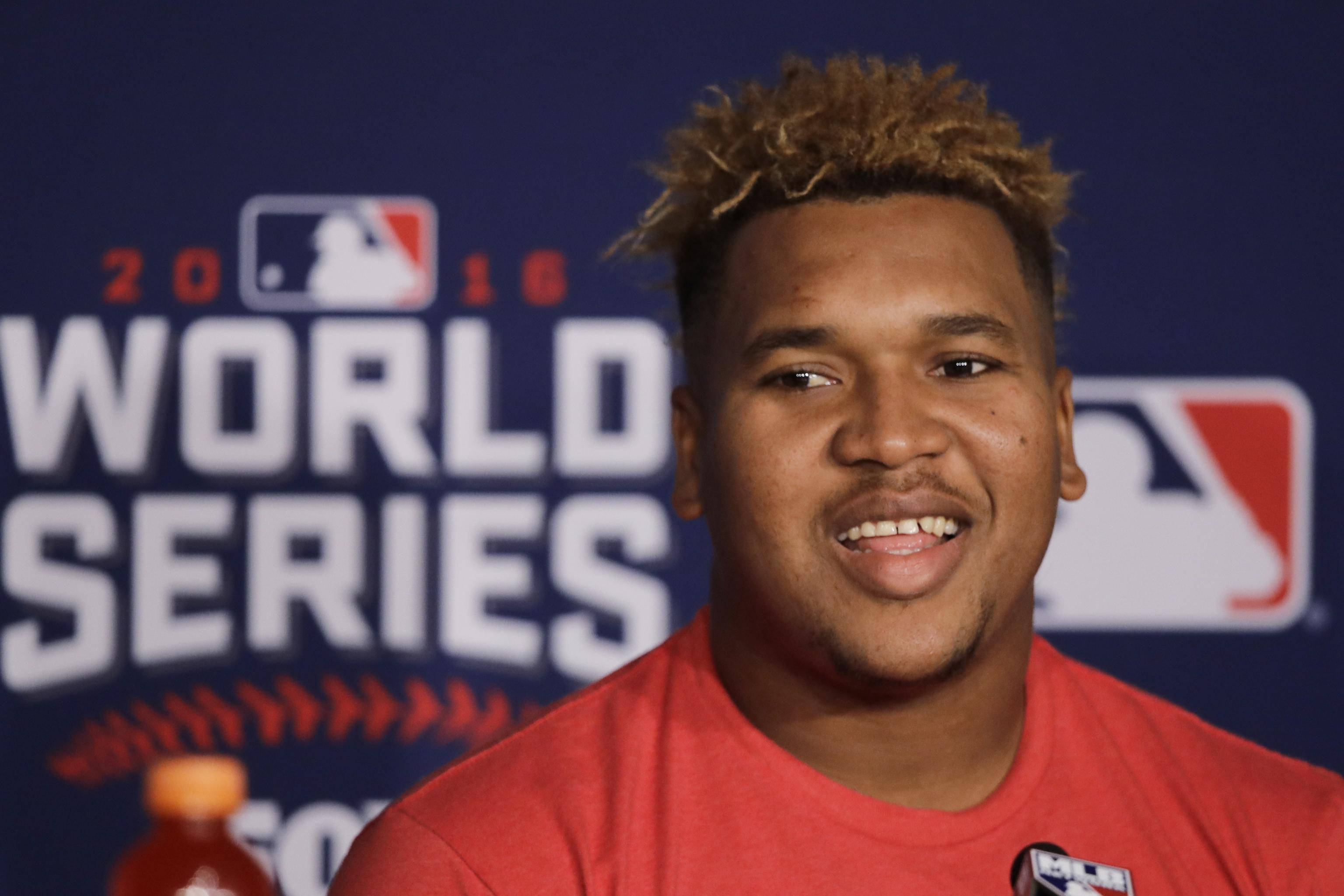 Jose Ramirez 短髮造型 壁紙 — 一位身穿紅色襯衫、面帶微笑的棒球運動員站在帶有MLB標誌的藍色2016年世界大賽背景前。