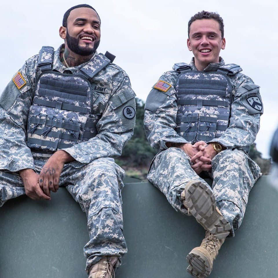 Joyner Lucas 於科切拉音樂節演出 壁紙 — 兩名面帶微笑的士兵穿著迷彩制服，在陰天戶外隨意坐在綠色屏障上。
