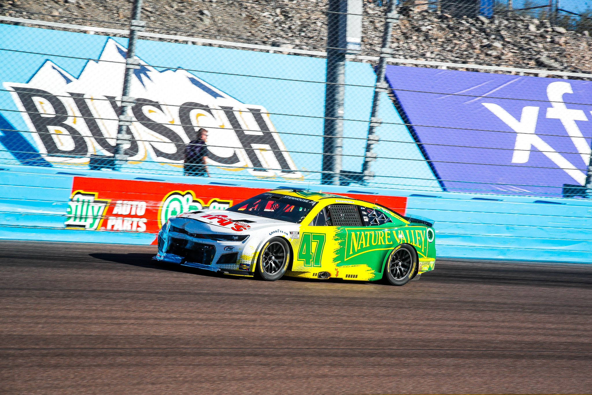 瑞奇·斯坦豪斯 Jr. 在賽道上疾馳 壁紙 — 一輛充滿活力的黃綠色NASCAR賽車在陽光明媚的賽道上飛馳，後面的藍色牆上有「Busch」和「Nature Valley」廣告。