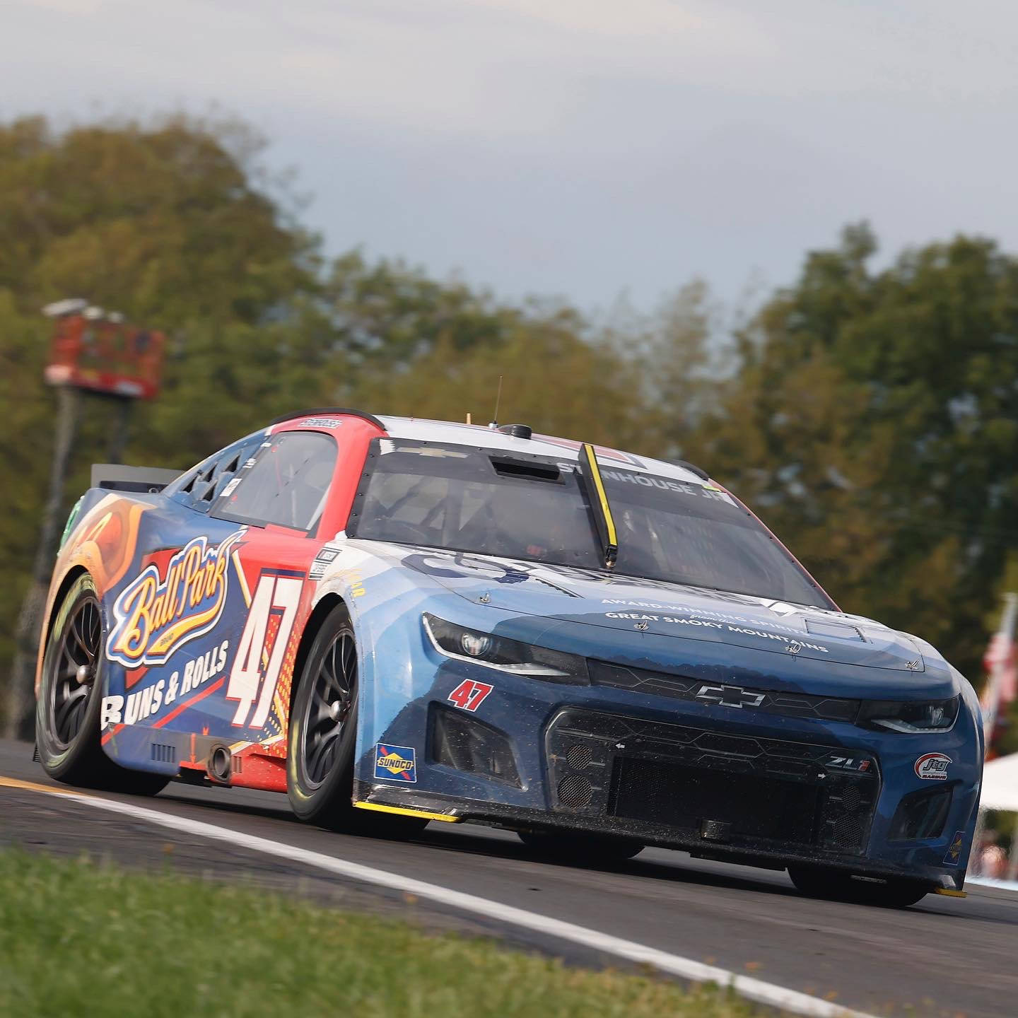 瑞奇·斯坦豪斯 Jr. 賽車特寫於賽道上 壁紙 — 一輛藍紅色的NASCAR賽車（47號）在陰天之下，沿著綠樹成蔭的賽道飛馳。