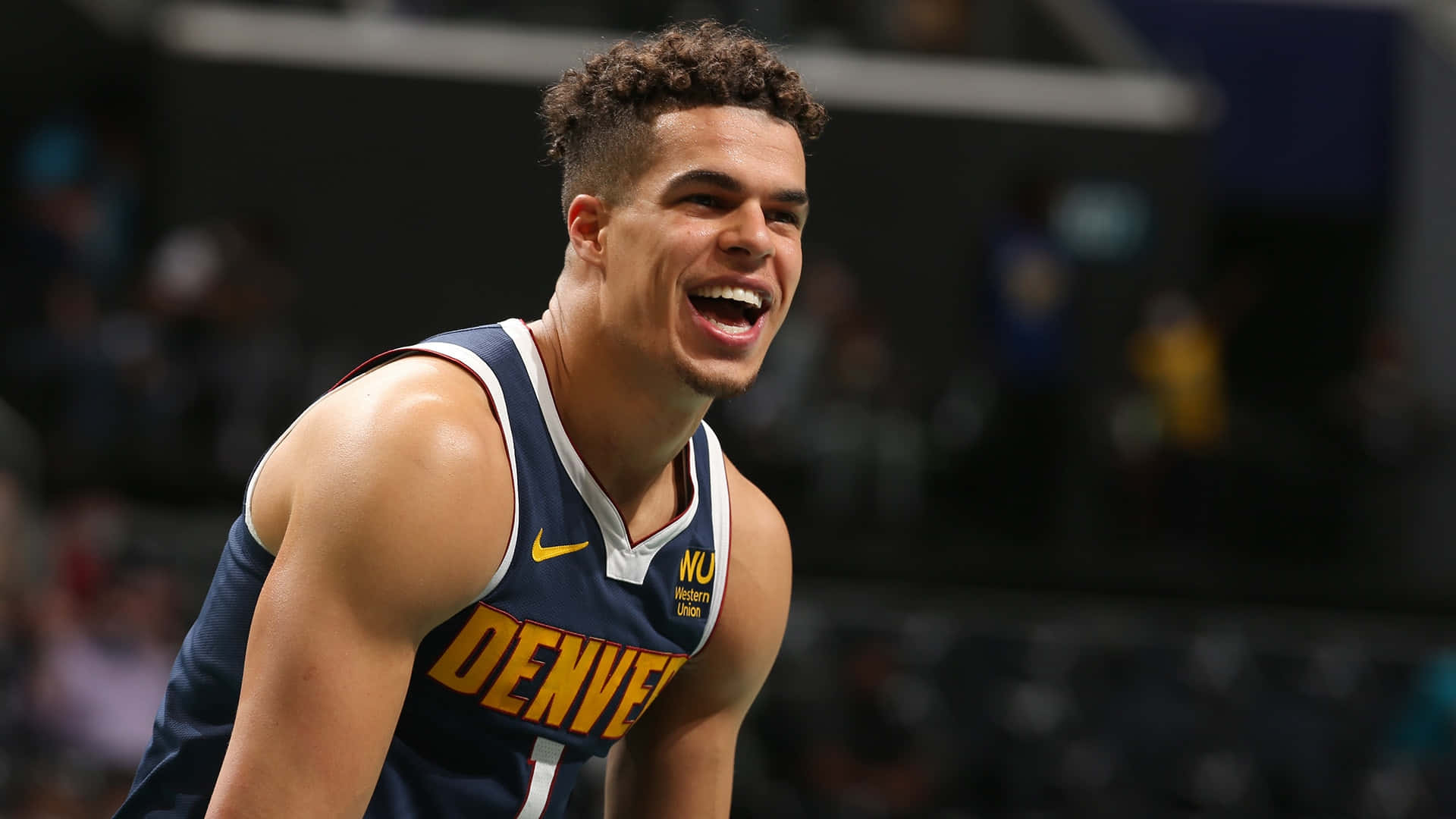 邁克爾·波特 Jr. NBA 丹佛金塊隊 壁紙 — 一名身穿丹佛金塊隊球衣的籃球運動員在球場燈光下比賽時開懷大笑。