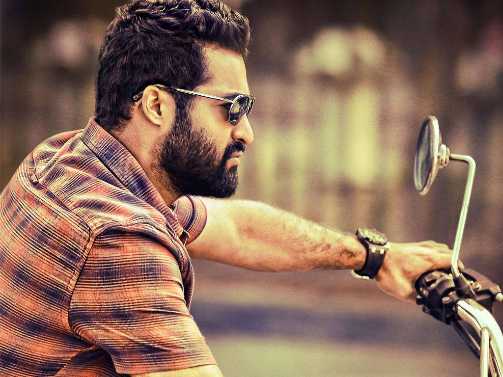 Jr Ntr 騎摩托車 壁紙 — 一個留鬍鬚的男人戴住太陽眼鏡，喺模糊嘅城市背景下，專注咁揸住電單車把手。