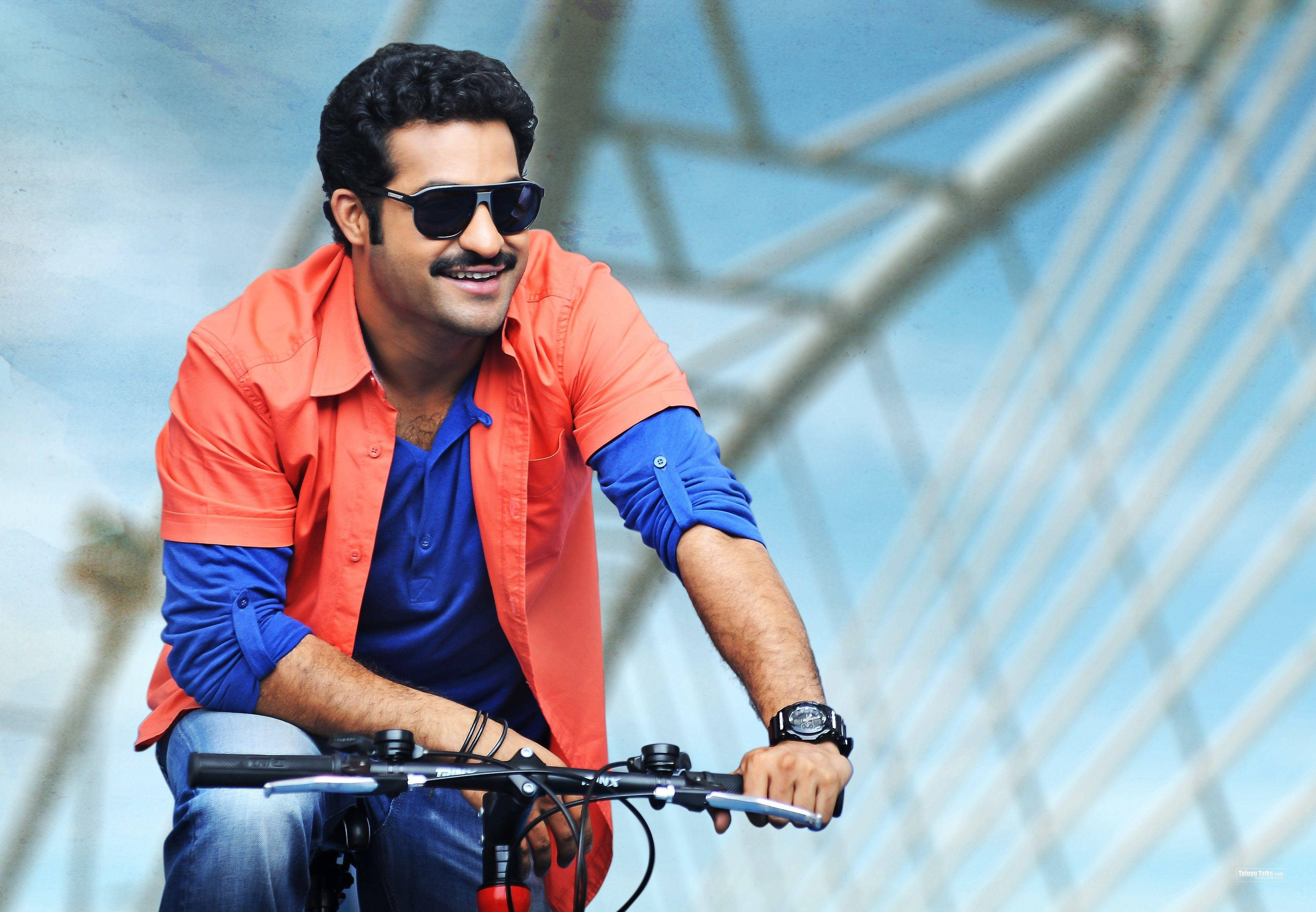 Jr Ntr 騎自行車 壁紙 — 一位微笑的男子戴著太陽眼鏡和彩色襯衫，在現代玻璃和鋼結構下騎自行車。