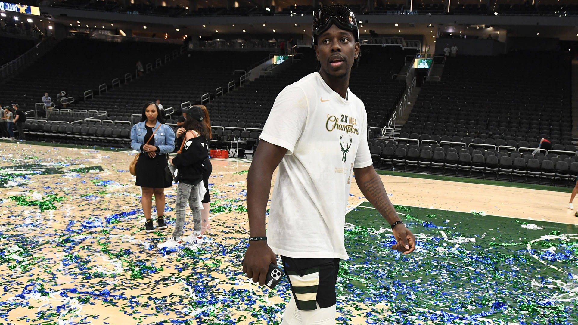 Jrue Holiday休閒裝扮 壁紙 — 一名身穿 '2x NBA Champions' 球衣的籃球運動員在空無一人的看台後方，走過灑滿彩紙的球場。