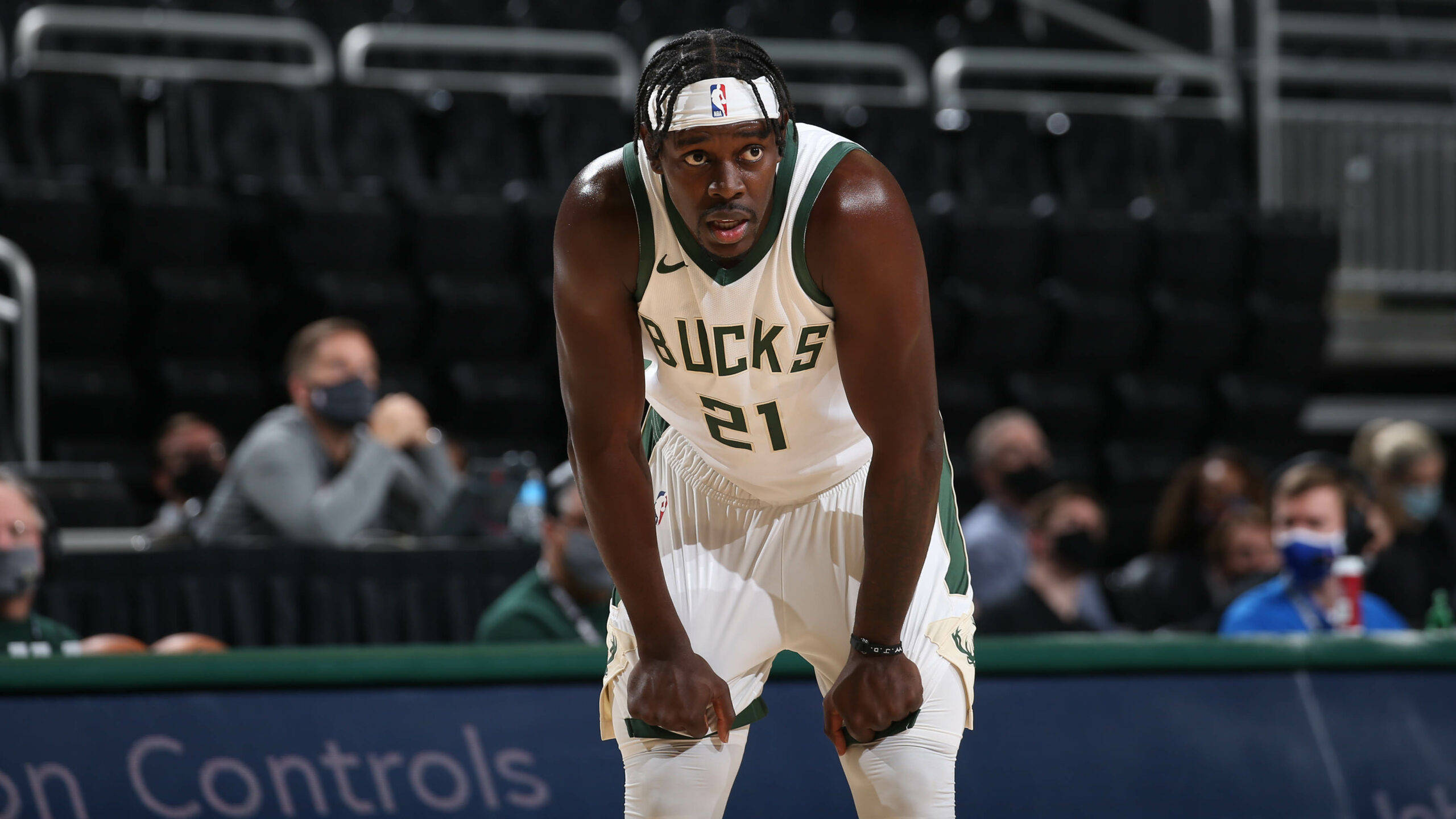 Jrue Holiday疲憊姿態 壁紙 — 一名身穿白色和綠色球衣的密爾沃基公鹿籃球運動員在球場上激烈前傾，背景有戴口罩的觀眾。