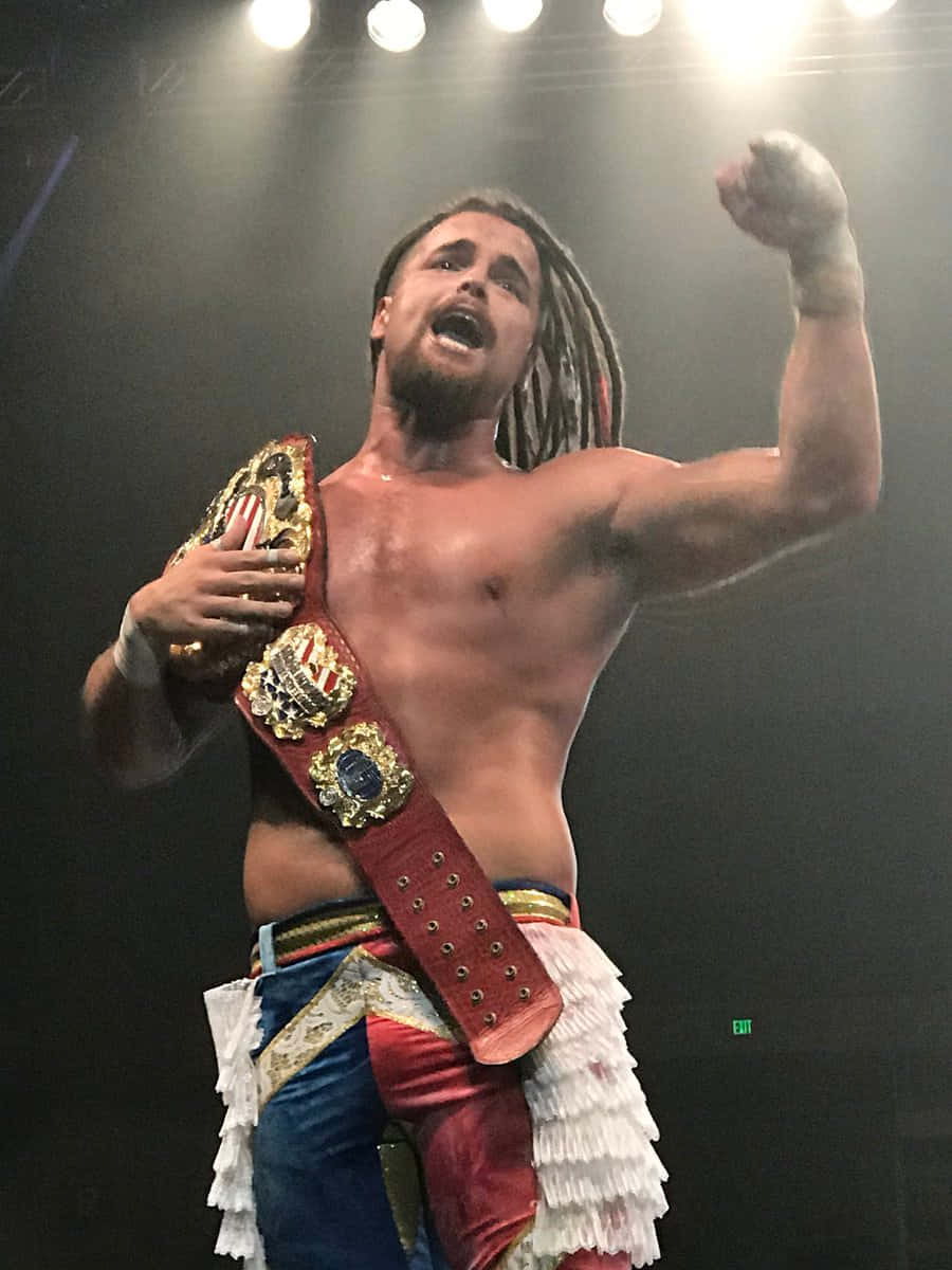 Juice Robinson 奪得冠軍腰帶 壁紙 — 一位赤膊、留著辮子的摔角手在明亮的舞台燈光下高舉冠軍腰帶，勝利慶祝。