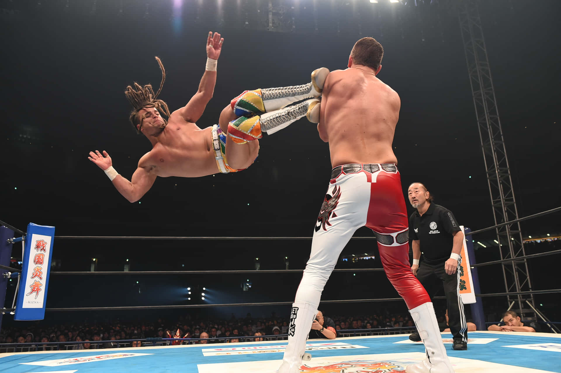 Juice Robinson 空中踢擊 壁紙 — 一名摔角手在半空中躍起使出高飛踢，對手在燈光明亮的擂台上做好準備，裁判在旁觀看。