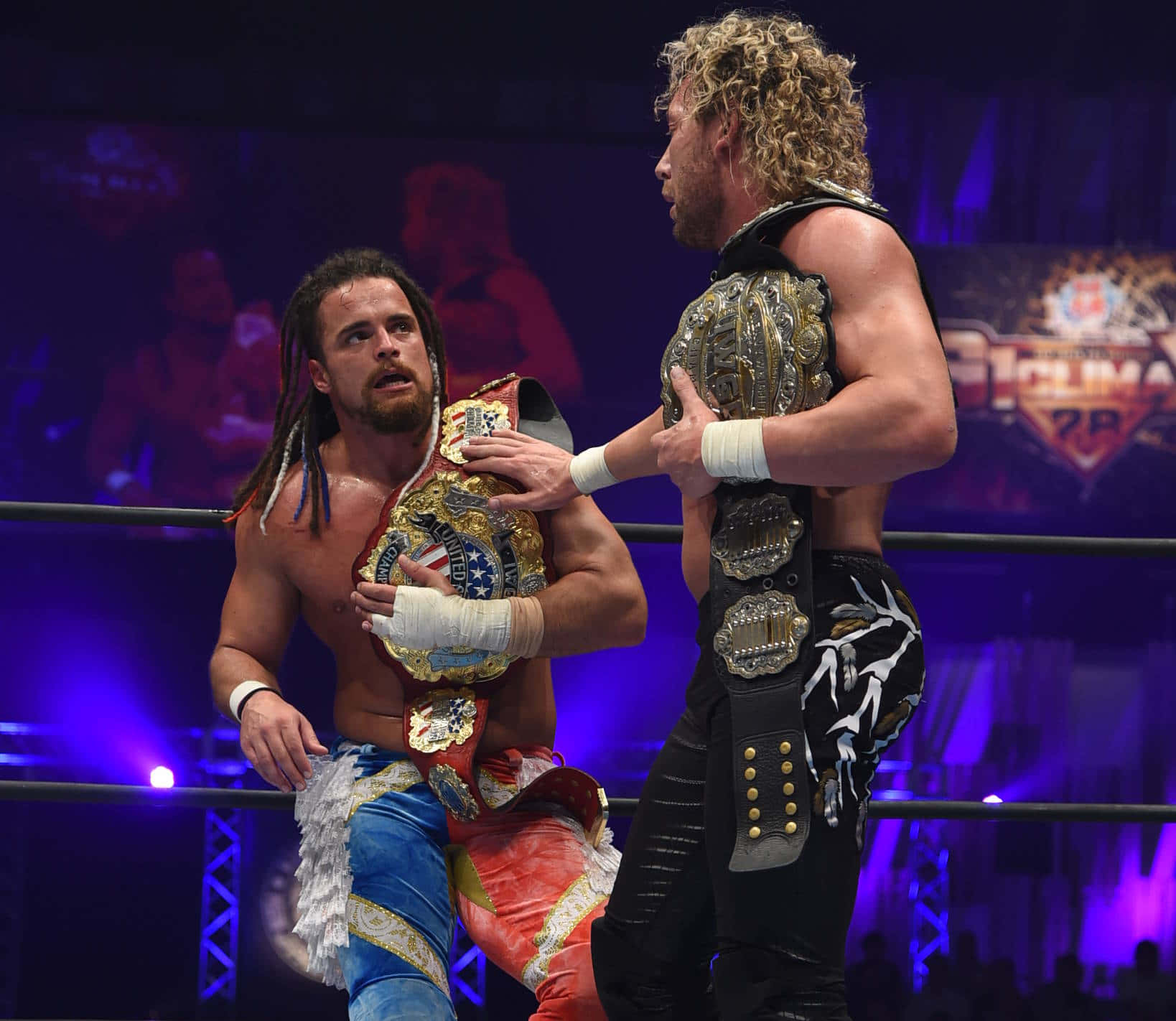 Juice Robinson 與Kenny Omega 壁紙 — 兩名肌肉發達的摔角手在擂台上激烈對峙，在明亮的舞台燈光下手持冠軍腰帶。
