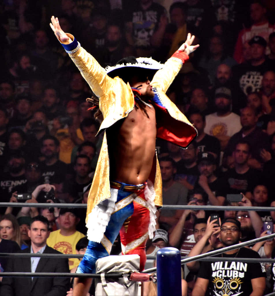 Juice Robinson 高舉雙臂 壁紙 — 一名赤裸上身的摔角手，身穿金色外套和戴上海盜帽，在歡呼的人群面前，雙臂高舉，勝利地站在擂台繩索上。