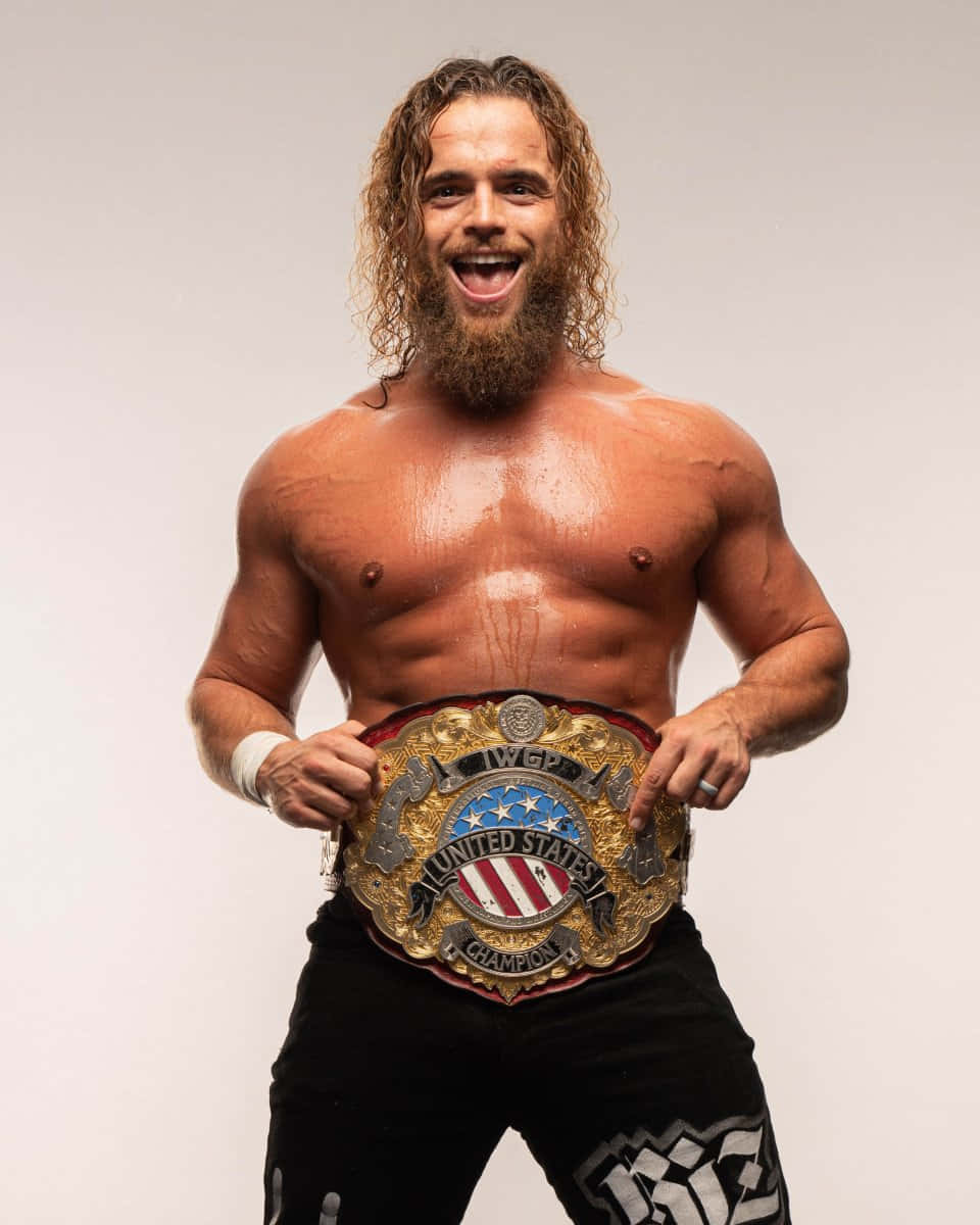 Juice Robinson 高舉冠軍腰帶 壁紙 — 一位汗流浹背、留著長髮的鬍鬚摔角手，在純色工作室背景前自豪地高舉IWGP美國冠軍腰帶。