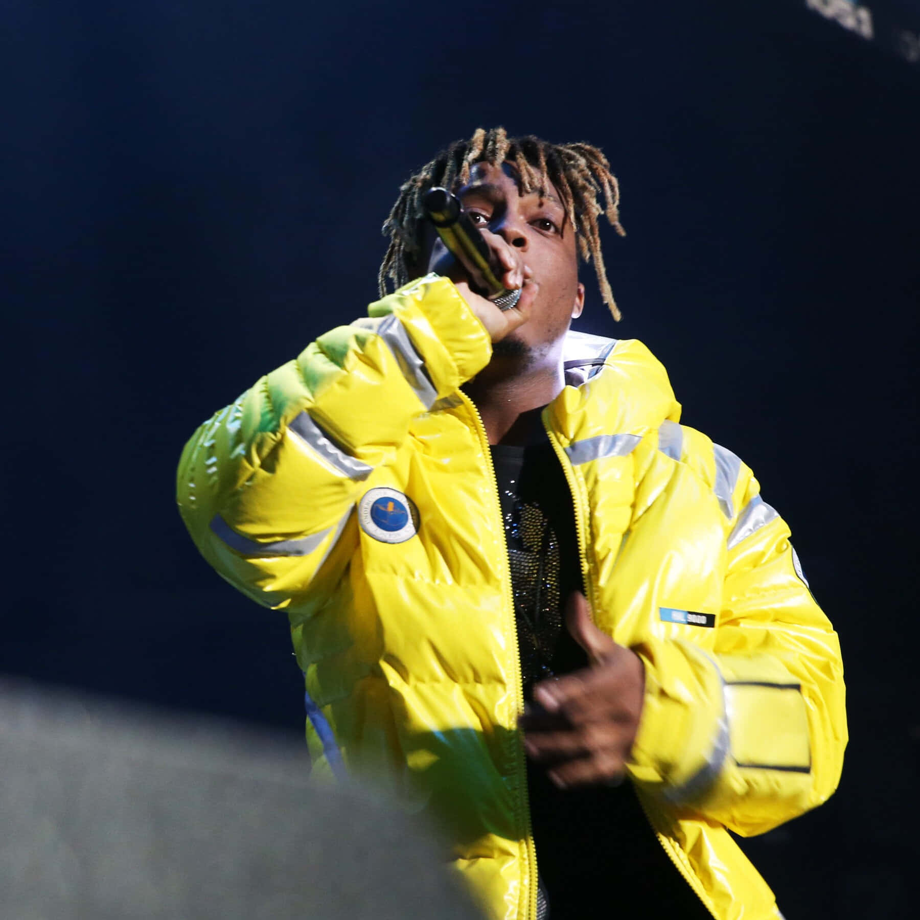Juice Wrld — 說唱先驅與開拓者 壁紙 — 一位梳著辮子的說唱歌手在昏暗燈光下穿著亮黃色羽絨外套在舞台上表演。
