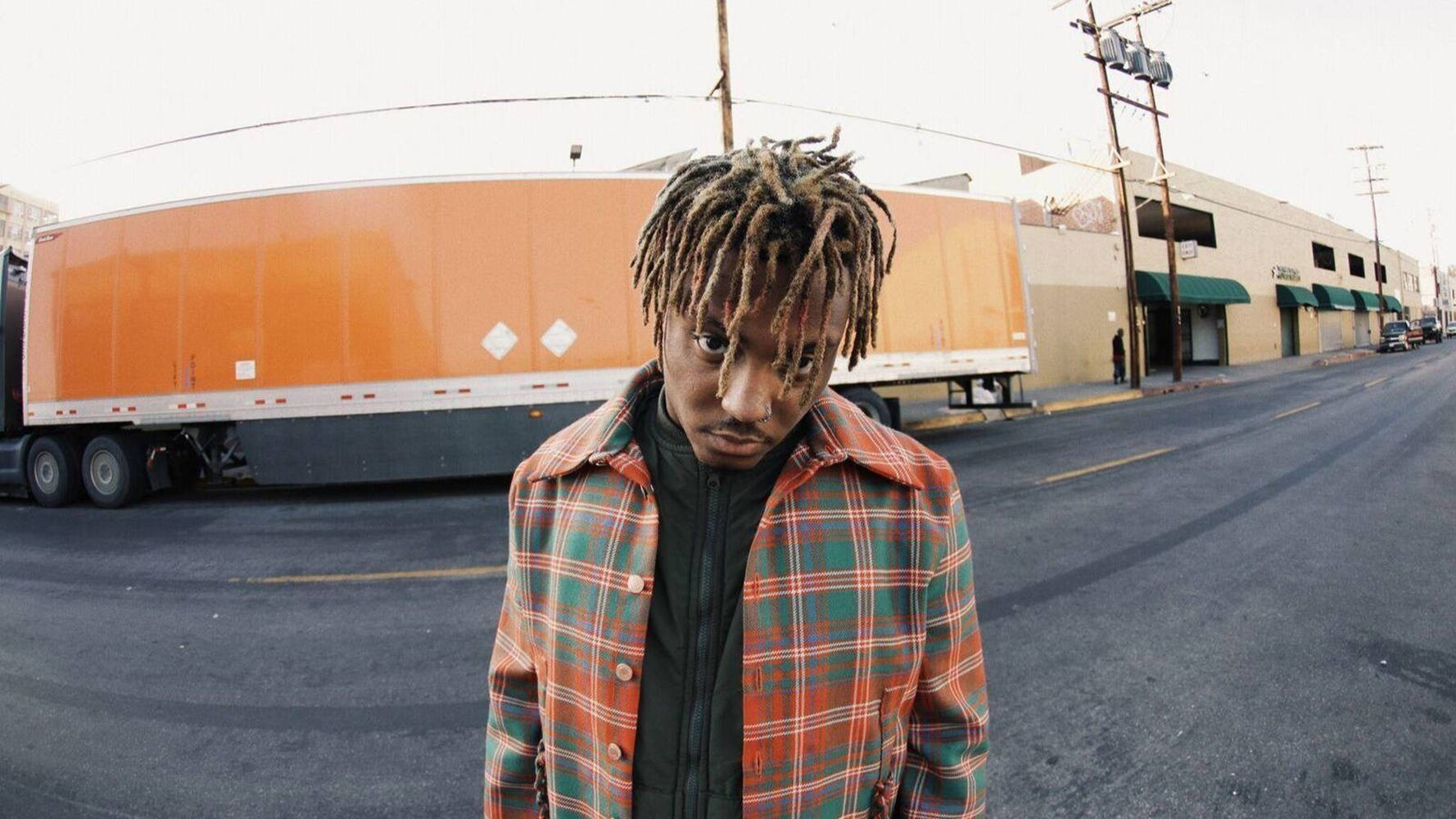 Juice Wrld 999 站在道路旁 壁紙 — 一位留著辮子的年輕男子在粗糙的城市街道上，旁邊有一輛橙色半掛車，他正凝視著鏡頭。