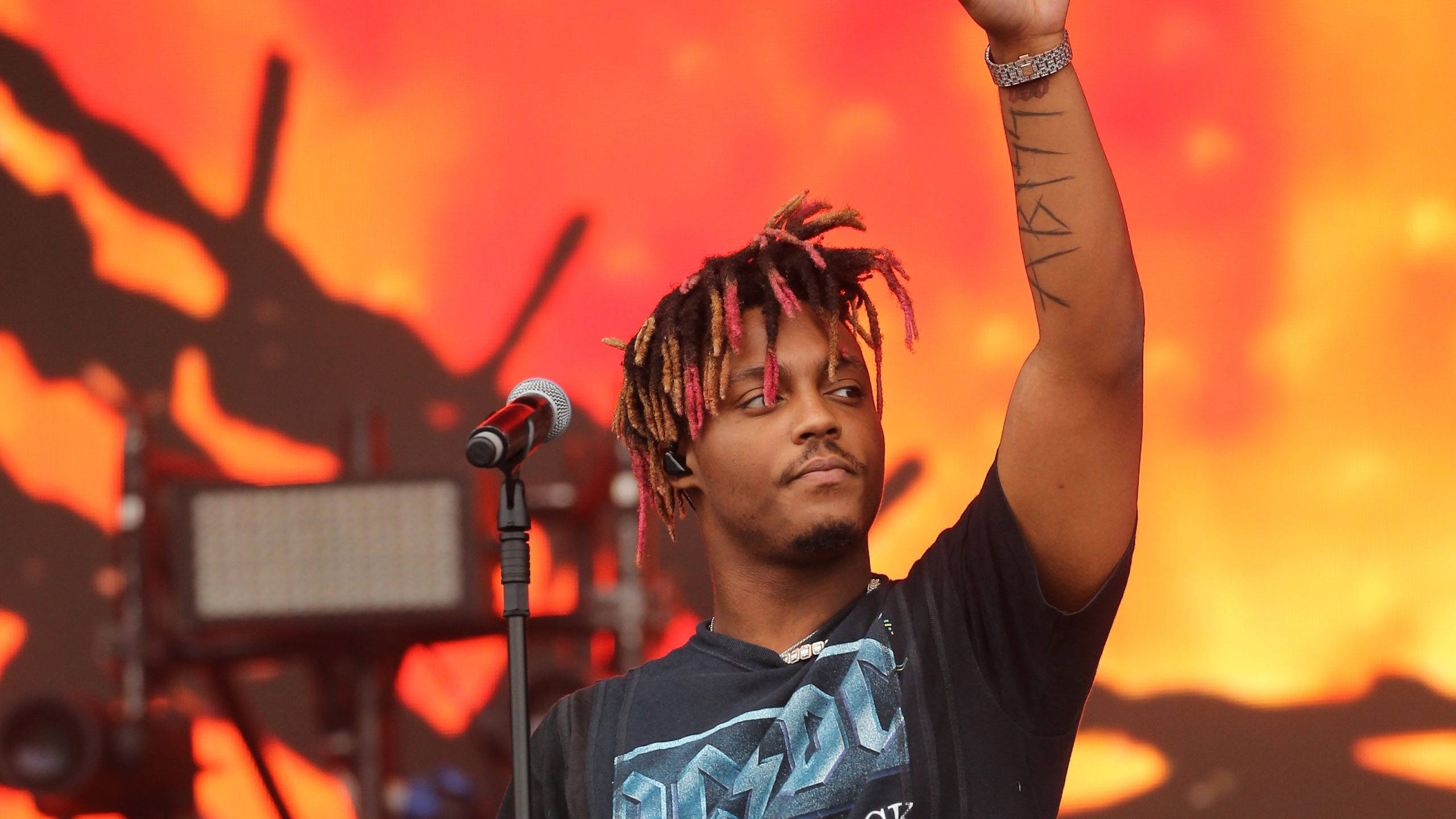 酷炫 Juice WRLD AC/DC 標誌演唱會 壁紙 — 一位男性表演者擁有色彩繽紛的辮子，在火橙色舞台背景上舉起他紋身的手臂。