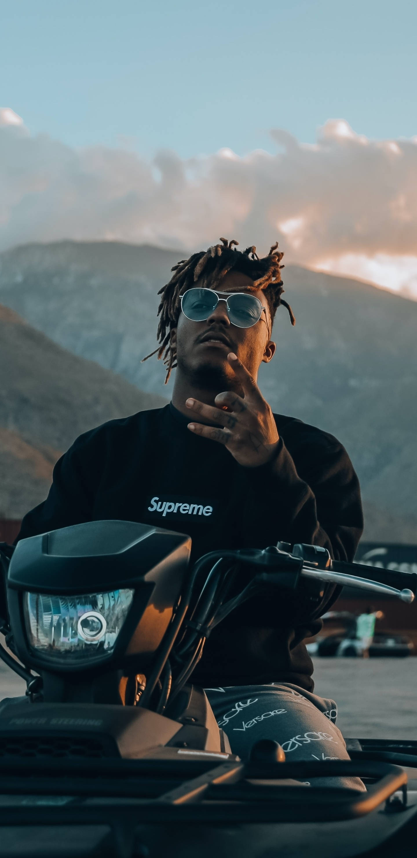 酷炫 Juice WRLD 騎 ATV 壁紙 — 一名留著辮子頭、戴著太陽眼鏡的男子，在日落時分自信地坐在摩托車上，背景是山巒。
