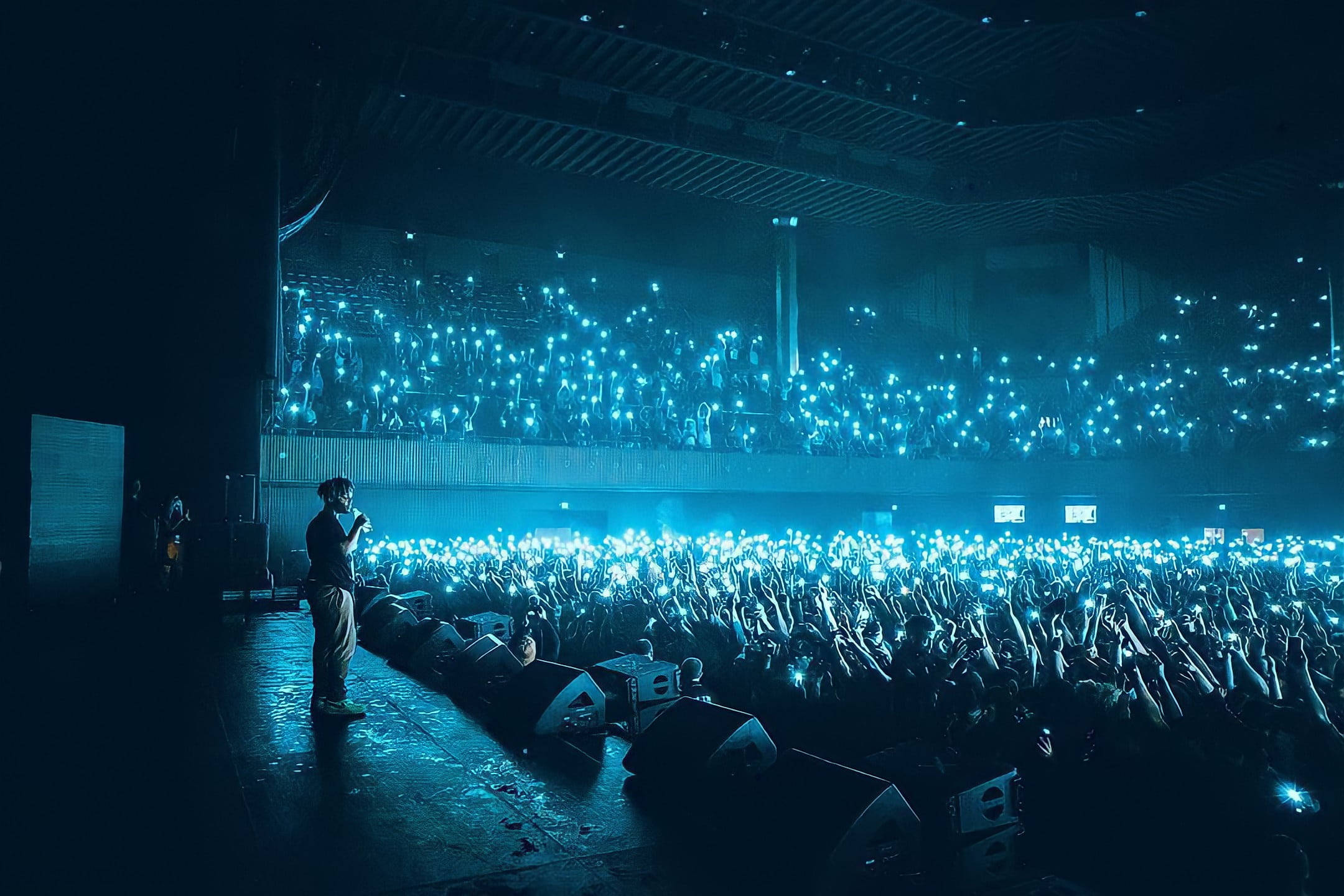 Juice Wrld 桌面壁紙 燈光 壁紙 — 一位表演者獨自站在黑暗的舞台上，沐浴在藍光中，而下面的粉絲海舉起發光的手機，創造出星光般的光波。